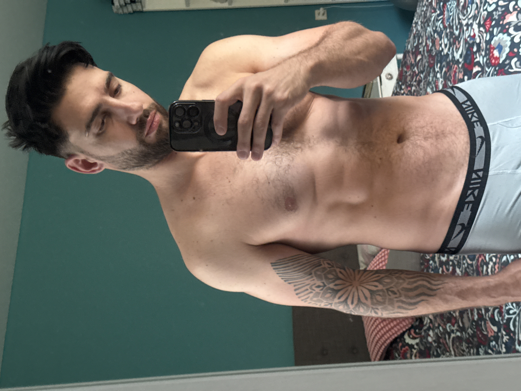 The image displays a mirror selfie of a man, presented sideways, captured from the waist up. The man has short, dark hair styled upwards and a neatly trimmed beard. He exhibits a muscular and fit physique, with visible definition in his arms and shoulders, and light body hair across his chest and abdomen. He is wearing light grey athletic underwear with a black and grey waistband that clearly displays the text "NIKE". A prominent and intricate tattoo, featuring mandala-like and wavy patterns, covers a significant portion of his left arm. He holds a dark smartphone, equipped with multiple rear cameras, in his right hand, looking towards it with a neutral and focused expression while taking the photograph.
The reflected background reveals a solid teal-green wall behind the man. To his right (viewer's left in the rotated image), a section of a bed is visible, covered by a vibrant, patterned duvet or comforter featuring red, white, blue, and black floral or paisley designs. A white trim or baseboard runs along the bottom of the wall. The lighting suggests an indoor setting, likely during daylight hours, within a private residence in Sofia, Bulgaria.