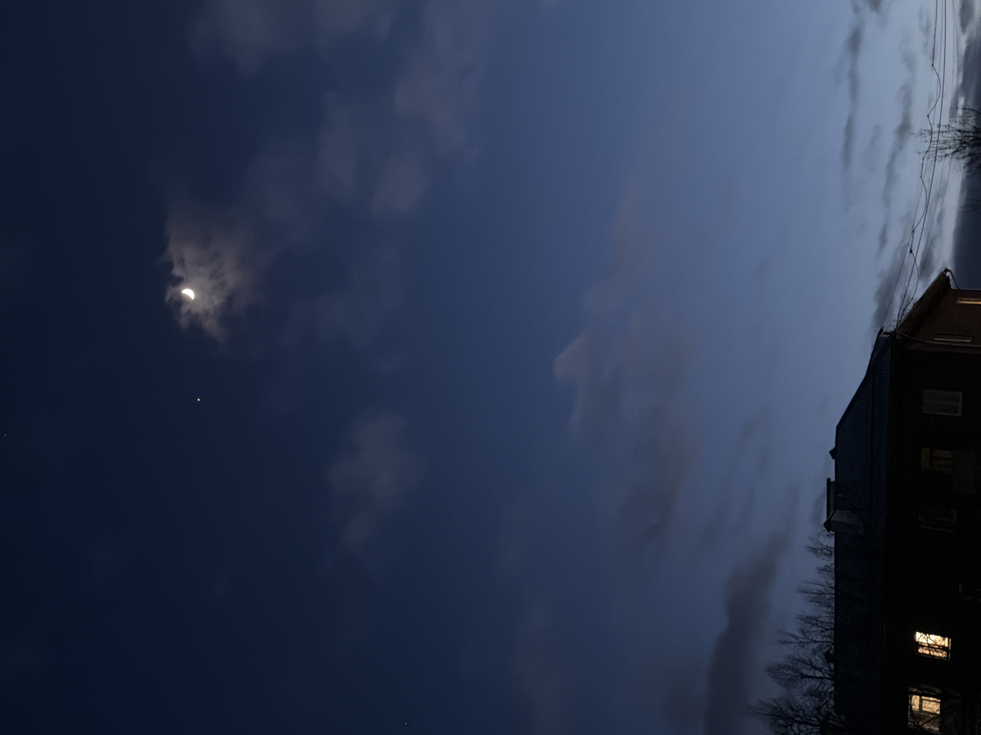 The image captures a serene nighttime or late evening scene dominated by a deep blue and grey sky. The perspective is tilted, but when viewed upright, the sky fills most of the frame.
In the upper left quadrant, a distinct crescent moon shines brightly through a break in the dark, wispy clouds. Below and slightly to the right of the moon, a single, brilliant yellow star or planet is visible against the dark sky. Clouds are scattered across the sky, varying in density from thin veils to darker, more substantial formations.
On the right side of the image, a dark, multi-story building, likely an apartment block or residential structure, is silhouetted against the sky. Warm, yellow lights glow from several windows on its facade, indicating activity within. Below and to the left of the building, the stark, bare branches of trees are visible, reaching up towards the sky. Overhead, a series of dark power lines stretch horizontally across the frame, suggesting an urban or suburban setting.
The overall mood is quiet and peaceful, characteristic of twilight or early night. The presence of the moon and a visible star indicates a relatively clear sky despite the clouds. The scene is set in Votkinsk, Russia. There are no people visible, nor is there any discernible text.