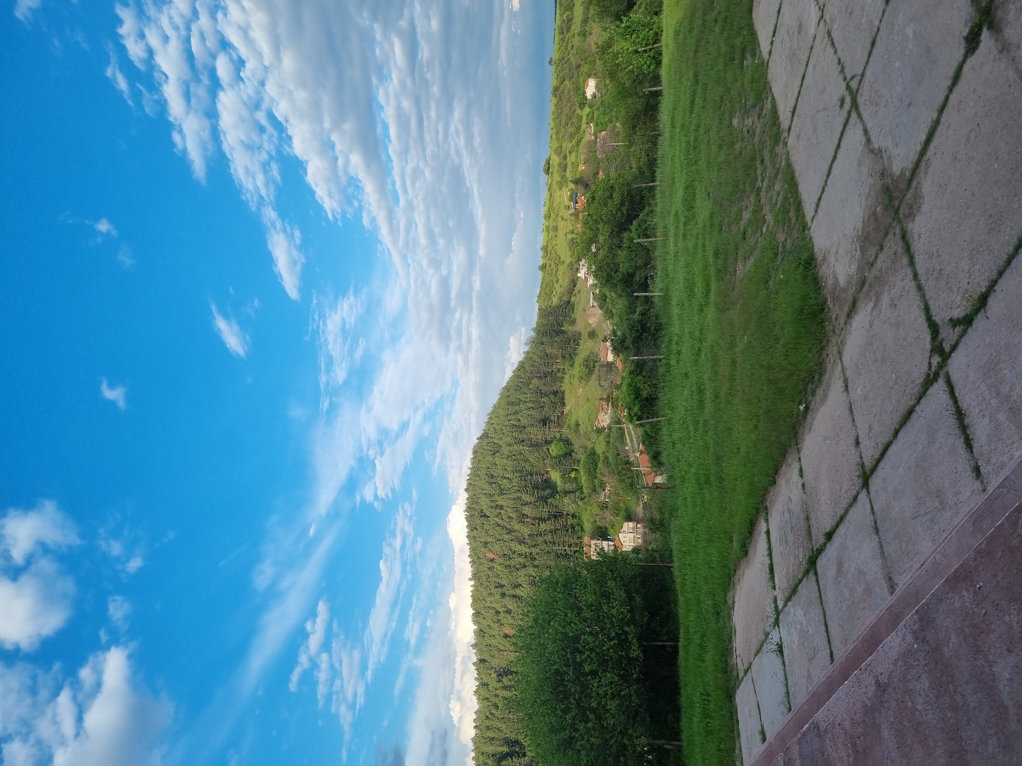 The image depicts a landscape in Gurgulyat, Bulgaria. The sky, predominantly blue with scattered white clouds, occupies the left portion of the frame. Adjacent to the sky is a tree-covered hillside dotted with houses. To the right of the hillside is a grassy area bordered by a paved walkway. The foreground includes a low, concrete wall.