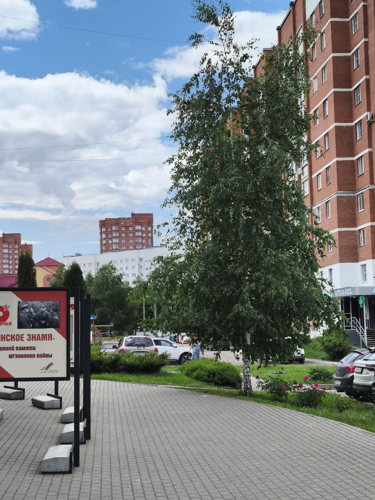 The image shows a paved area in front of a modern red brick apartment building. A tall, leafy birch tree stands in front of the building, obscuring part of its facade. To the left of the tree, a portion of a display board is visible. It has a red border and appears to feature an image of people in military uniforms and text in Russian, including the words "МАЯ" and "НСКОЕ ЗНАМЯ." Further to the left, a large red star symbol is partially visible on the display board. In the background, other apartment buildings of similar architectural style are visible. Several cars are parked on a grassy area next to the paved walkway. A few people can be seen walking in the distance. The sky is blue with scattered white clouds. The scene appears to be a public space within a residential area of Малая Валяевка, Russia, possibly near a monument or memorial given the content on the display board.