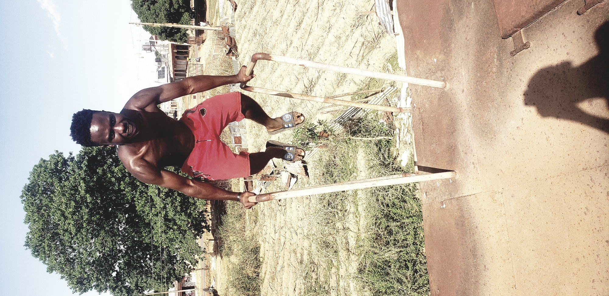 A shirtless man in red shorts and sandals is performing a handstand on parallel bars. The scene is in an outdoor location, possibly Narayi, Nigeria, given the request. The ground is dry, grassy and brown, with some scattered debris. A large tree is to the left. Behind the man, there is a building in the distance. To the right, there is a large, rusty metal structure. The shadow of an individual taking the photograph is visible on the metal structure.