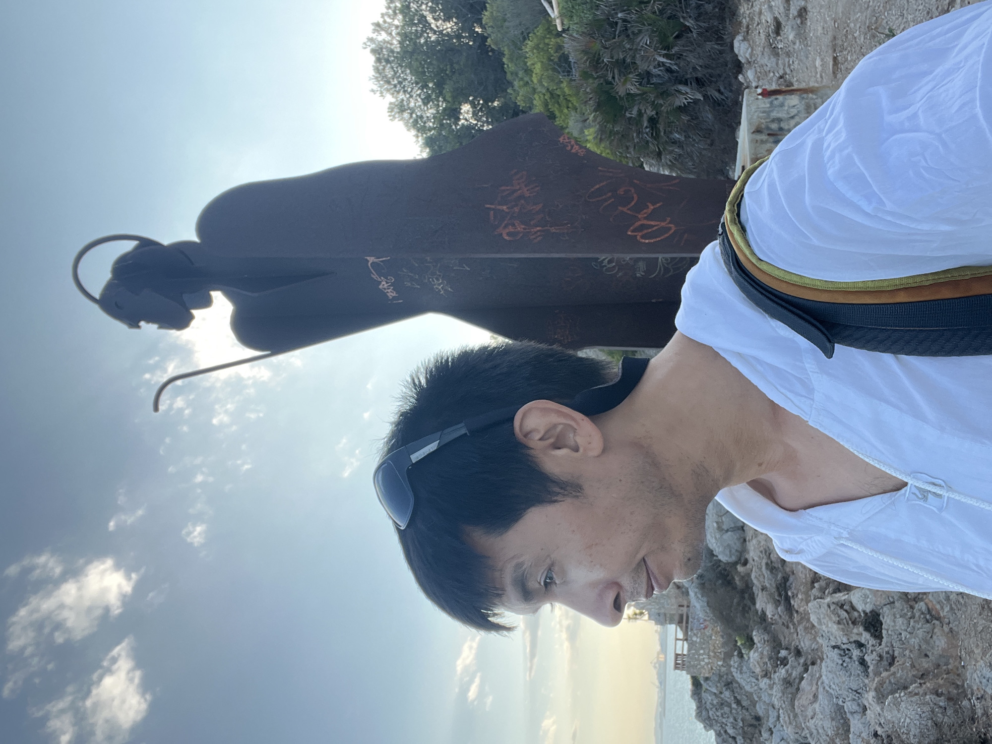 A man, facing left in profile, is in the foreground of an outdoor scene. He has short, dark hair, wears sunglasses perched on his head, and is looking towards the left. He is wearing a white hooded shirt and a bag strap is visible over his right shoulder. Behind him, to his right, is a tall, dark, metal statue of a figure with a halo-like structure above its head and a staff. The statue has orange graffiti on its surface. The background shows a rocky coastline with the sea and distant structures visible on the horizon. Lush green vegetation is present to the right of the statue. The sky is a pale blue with scattered white clouds and the sun is shining, casting a warm light on the scene. The location is identified as El Miracle, Spain.