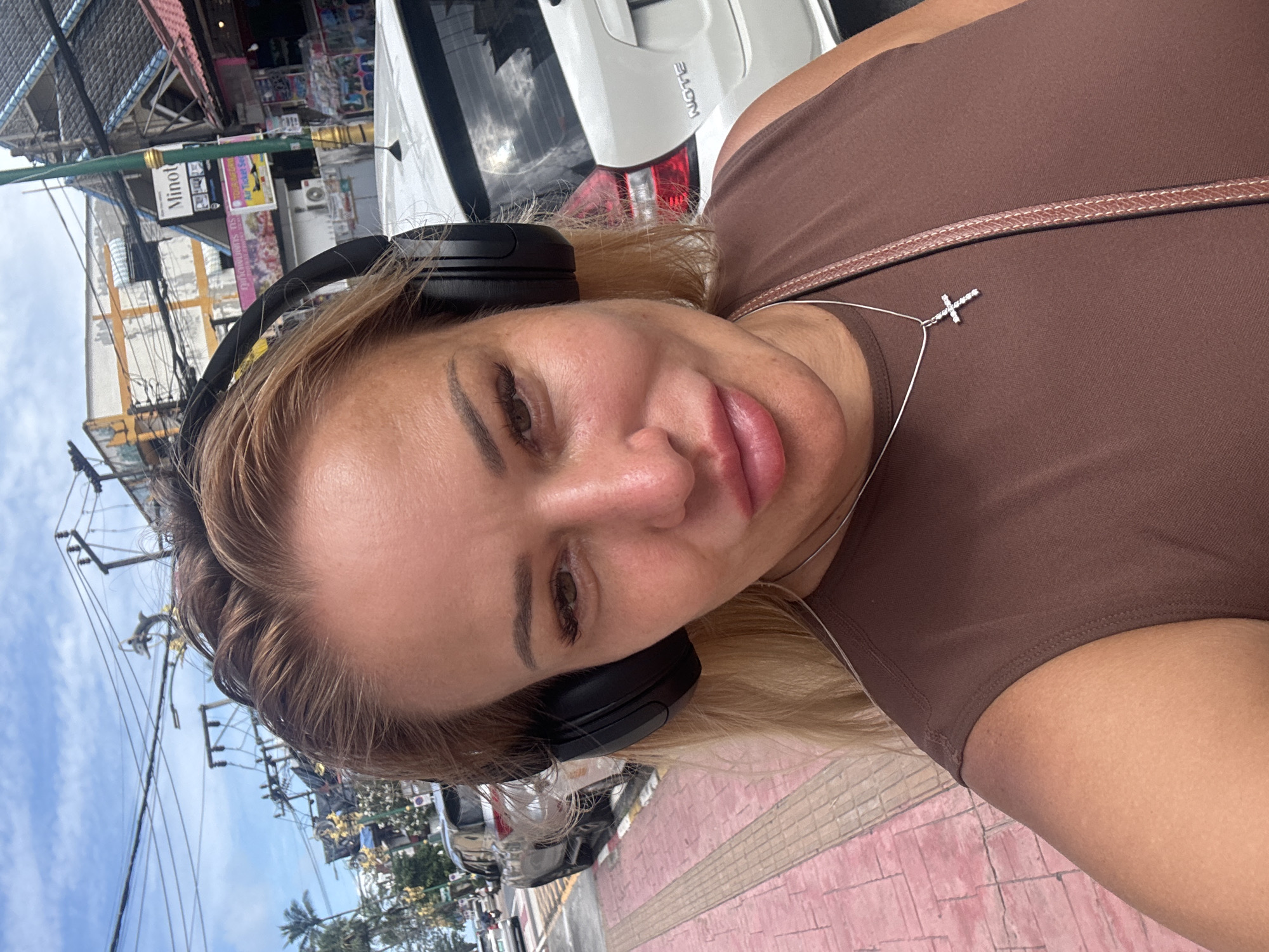 A woman with blonde hair, wearing black headphones and a brown tank top, takes a selfie outdoors. She is adorned with a silver necklace featuring a cross pendant. The background reveals a street scene in Patong, Thailand, with buildings, power lines, parked cars, and a sidewalk. The sky above is partly cloudy.