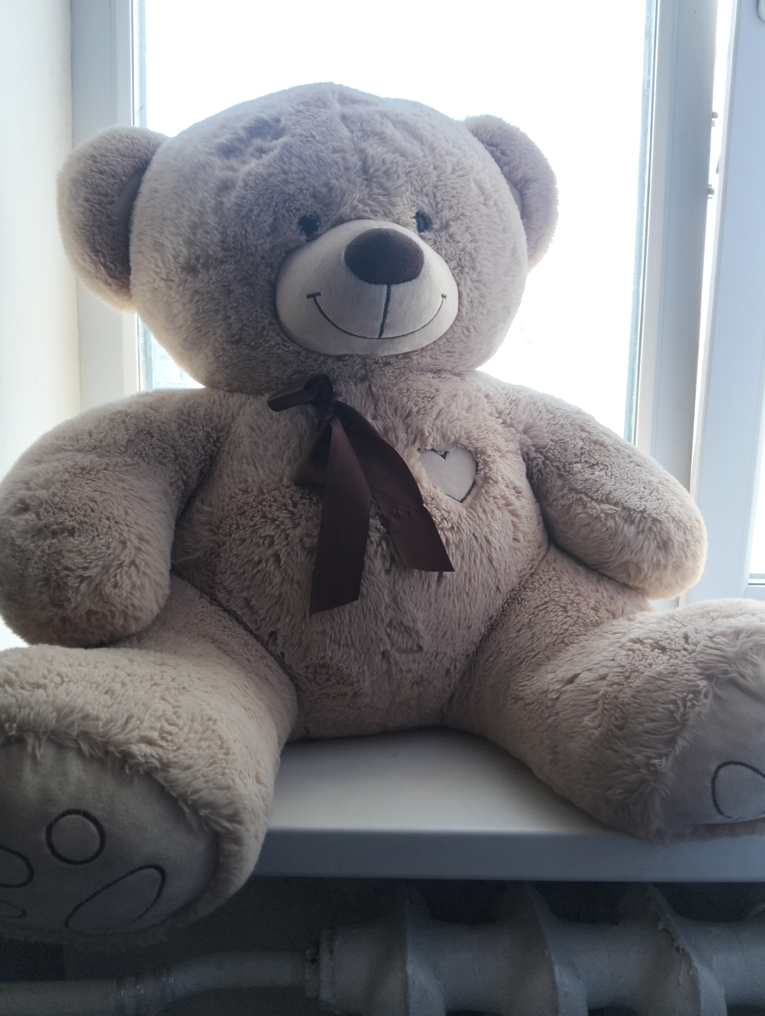 A large, plush, light brown teddy bear sits on a windowsill. The teddy bear is facing the viewer and has a brown ribbon tied in a bow around its neck. A small heart shape is embroidered on its chest. The background shows a bright window, suggesting it is daytime. Below the windowsill, a radiator is visible. The image is a photograph taken in the city of Cherkessk, Russia.