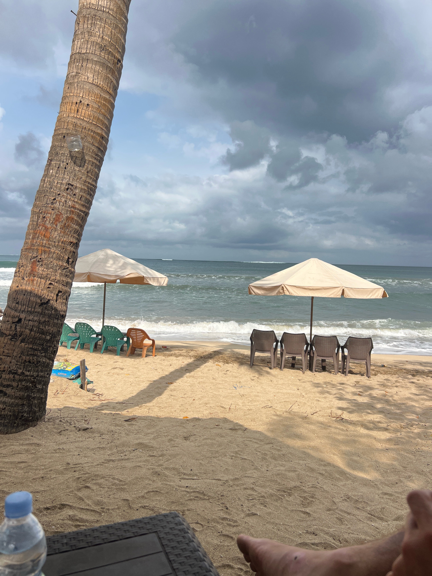 The image depicts a beach scene in Kuta, Indonesia, under a cloudy sky. In the foreground, a large palm tree trunk is visible on the left, with a discarded plastic bottle attached to it. A woven table with a bottle of water on it is positioned in the lower left corner. The sand stretches towards the ocean, with two beach umbrellas providing shade. Under the left umbrella, several green plastic chairs and one orange plastic chair are arranged, along with a blue inflatable object on the sand. Under the right umbrella, four brown plastic chairs are lined up. The ocean has moderate waves, and the sky is filled with dark, ominous clouds, suggesting the possibility of rain. The overall impression is one of a quiet beach, possibly before or during a storm. A person's feet are visible in the lower right corner of the frame, indicating someone is relaxing on the beach.