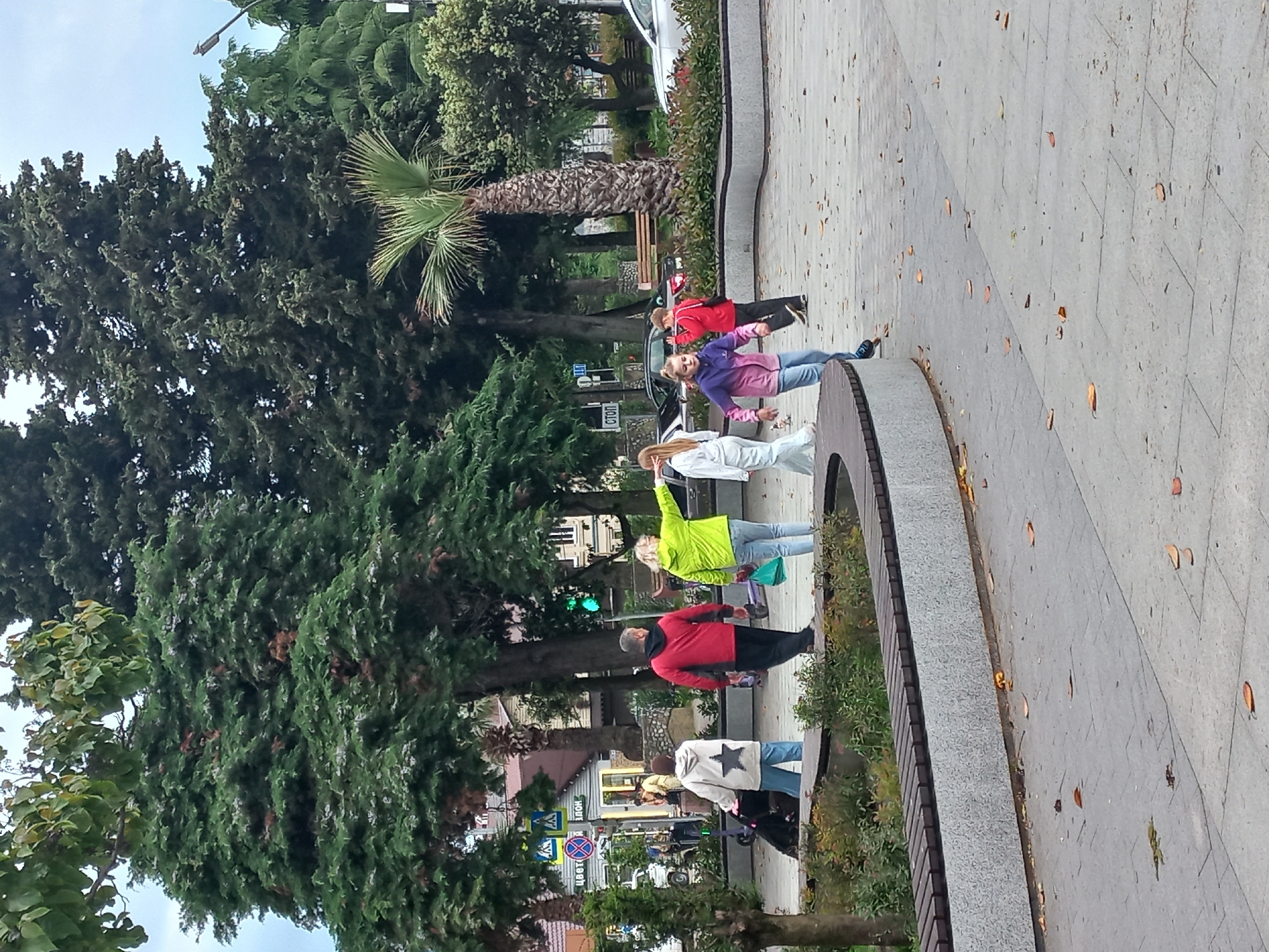 The image shows a street scene in Sochi, Russia, with a group of people walking along a sidewalk bordered by trees and greenery. The sidewalk is paved with gray stones and scattered with leaves. A row of people, five in total, are walking along the sidewalk. From left to right, a person in a white jacket with a star, a person in a red jacket, a person in a green jacket with their arm raised, a person in a white jacket, and a person in a purple jacket. The background includes a variety of trees, including palm trees, and buildings across the street. A white car is parked in the background, and street signs are visible. The environment suggests an urban park or recreational area.