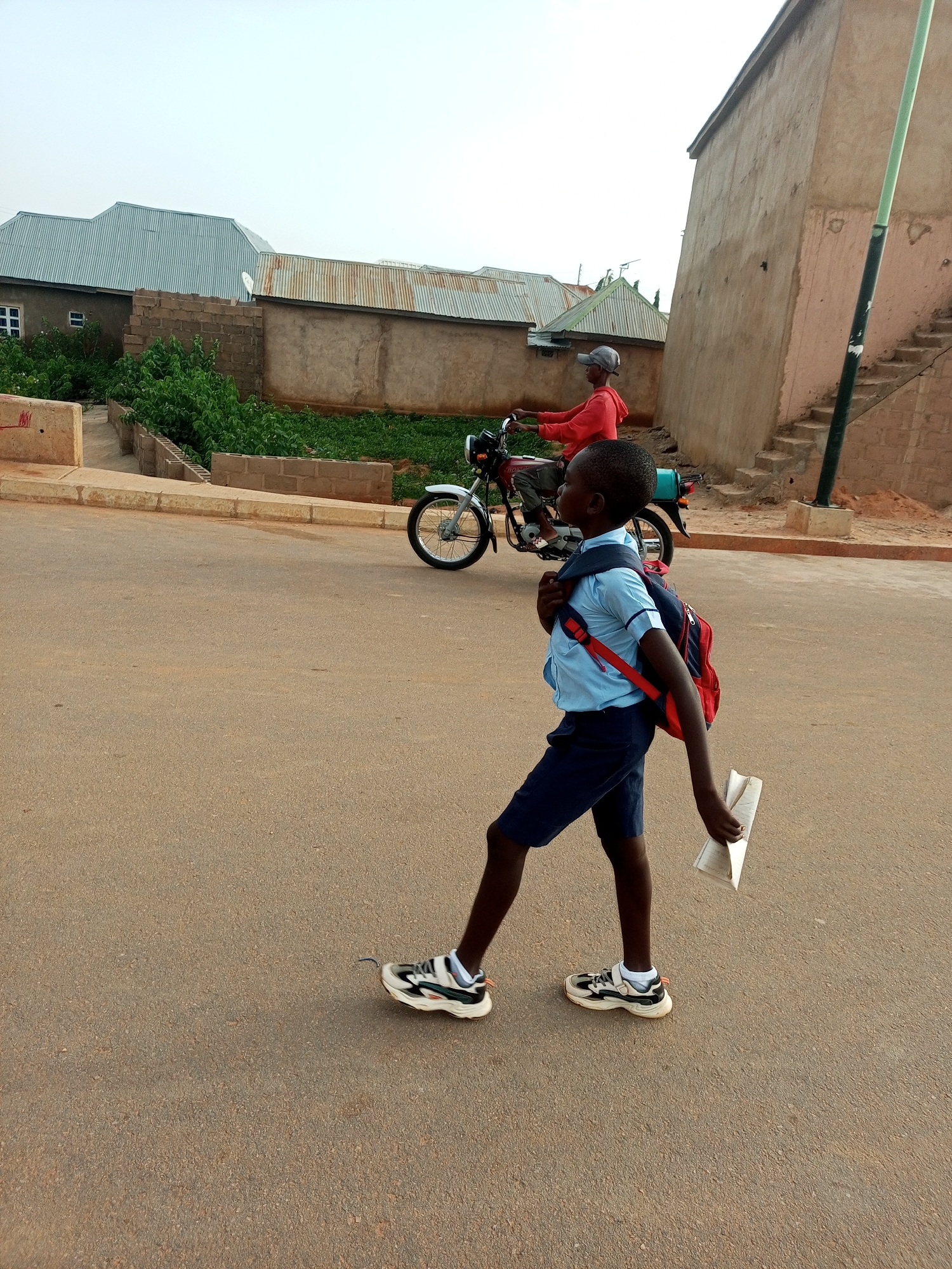 The image depicts a street scene in Tempure, Nigeria. In the foreground, a young boy wearing a light blue short-sleeved shirt, dark blue shorts, white socks, and black and white athletic shoes walks on a paved road. He carries a red and blue backpack and holds a folded paper document in his right hand. Behind him, a person wearing a red long-sleeved shirt and a grey baseball cap rides a maroon motorcycle. The background reveals residential buildings with corrugated metal roofs and some overgrown vegetation. A concrete structure with visible stairs is also present on the right side of the image. The sky is overcast.