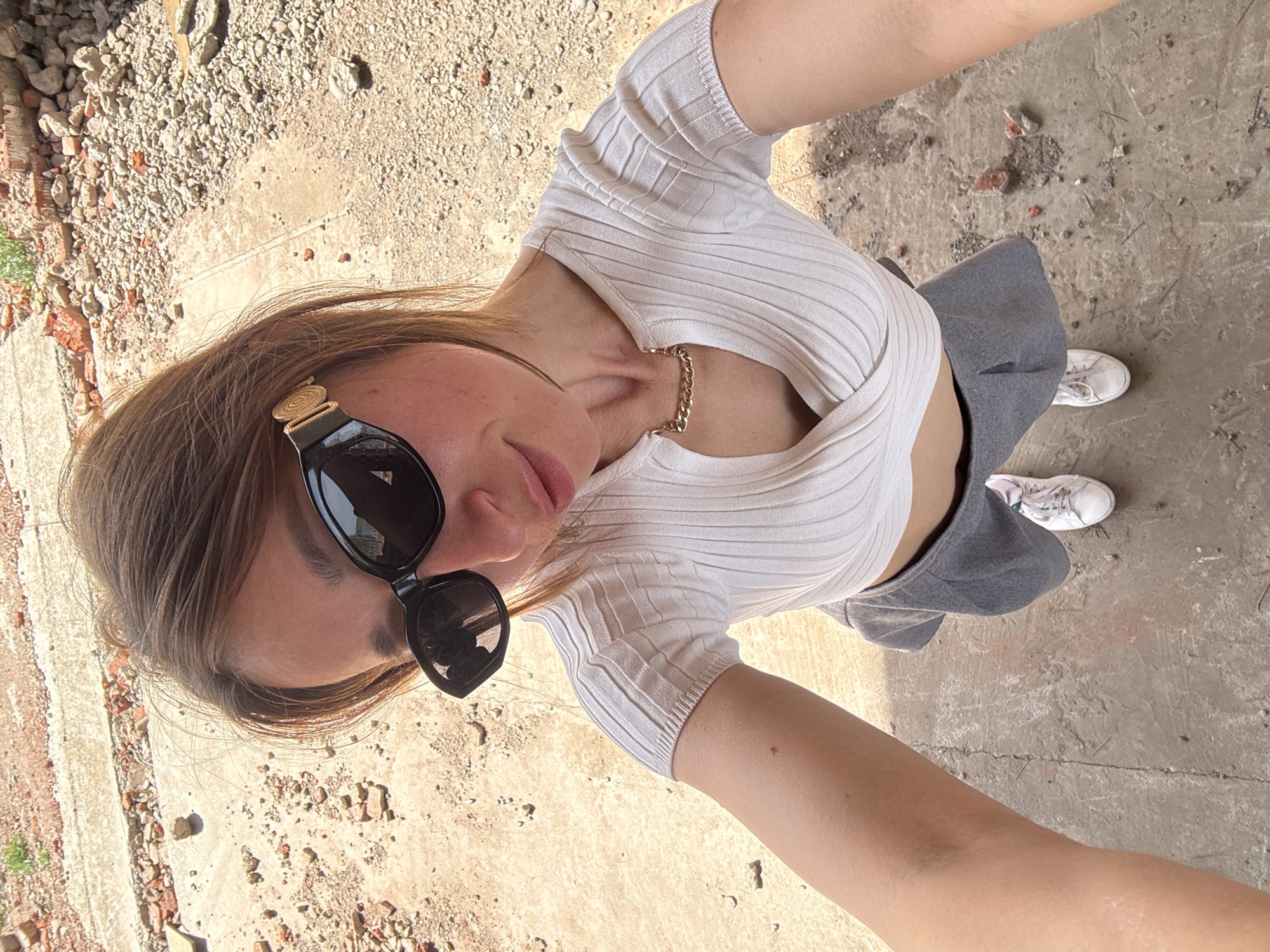 A young woman stands on a concrete surface with rubble and debris, possibly in an abandoned or dilapidated area. She is wearing a white, form-fitting, short-sleeved shirt, a gray skirt, and white sneakers. Sunglasses with gold-toned detailing on the temples are perched on her face. She has light brown hair and a gold chain around her neck. The angle of the photo suggests she is holding the camera herself, taking a selfie from a high vantage point. The location appears to be an outdoor area with the concrete surface cracked and broken in places, and with scattered brick and rubble. The location is in the city of Chaikovsky, Russia.
