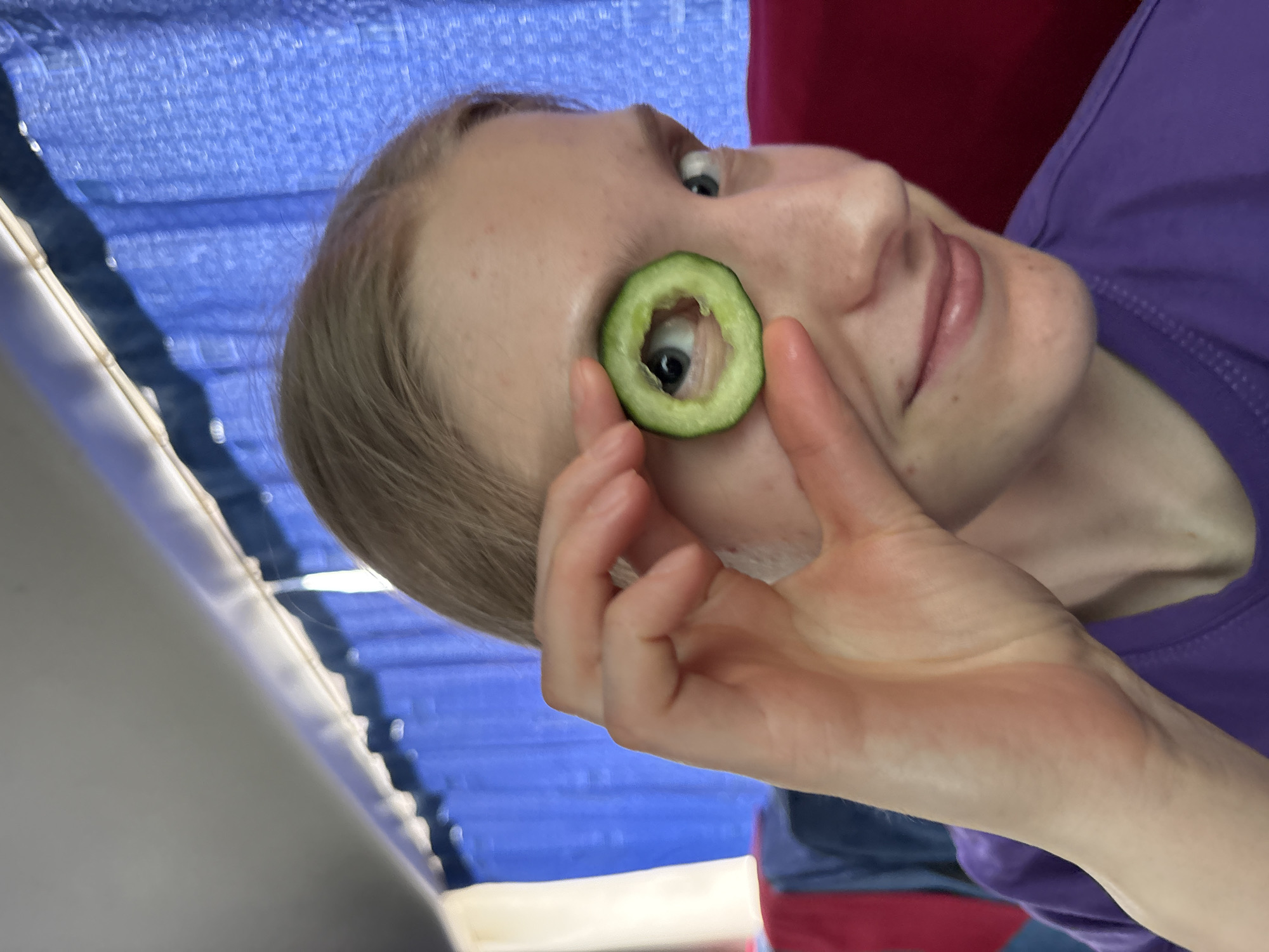 The image shows a fair-skinned person with blonde hair holding a slice of cucumber in front of their left eye. The person is wearing a purple shirt. The background includes blue fabric and a red surface. The location in the city of Yanychi, Russia, cannot be determined from the information provided in the image.