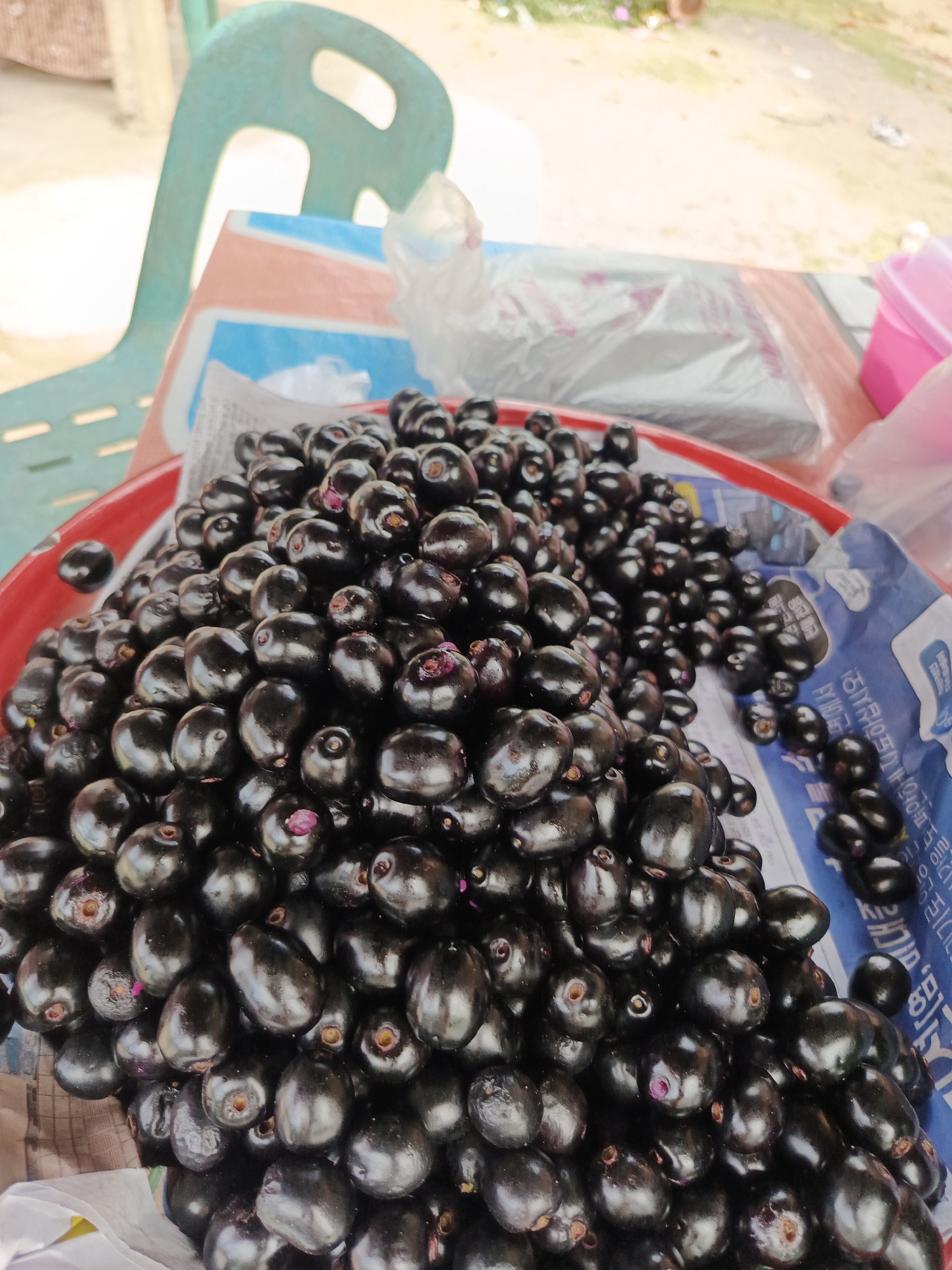 The image shows a close-up of a pile of ripe jamun fruits (also known as Java plum or black plum) in a red plastic bowl. The fruits are dark purple to black, oval-shaped, and glossy. Some fruits have small purple remnants of their stems. The bowl is placed on a table with a newspaper underneath it and a plastic sheet partially covering the table. In the background, a portion of a teal-colored plastic chair and some out-of-focus vegetation and structures are visible. The scene appears to be an outdoor market or roadside stall, likely in Sawang, Indonesia, given the context provided. The lighting suggests it is daytime.