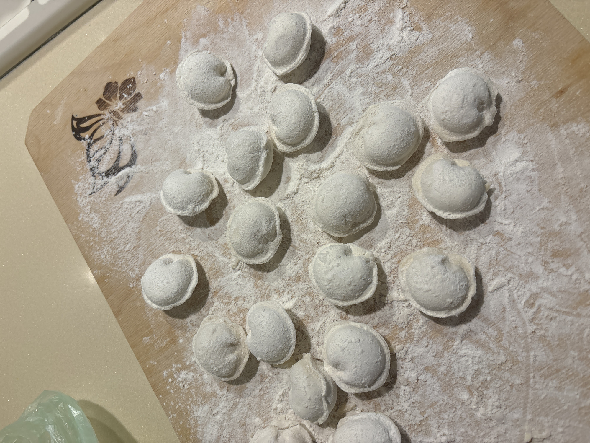 The scene, located in the city of Chaikovsky, Russia, displays approximately twenty-five uncooked pelmeni (dumplings) scattered across a wooden cutting board. The board is extensively dusted with white flour, and a dark, stylized floral or butterfly pattern is visible on its upper right, partially obscured by flour. The pelmeni are light in color, coated in flour, and exhibit typical crimped edges. A light beige countertop is visible along the top and left edges of the cutting board, with a crumpled, translucent light green plastic bag partially visible in the upper left corner. No individuals are present in the frame. The environment indicates a domestic kitchen setting during food preparation.