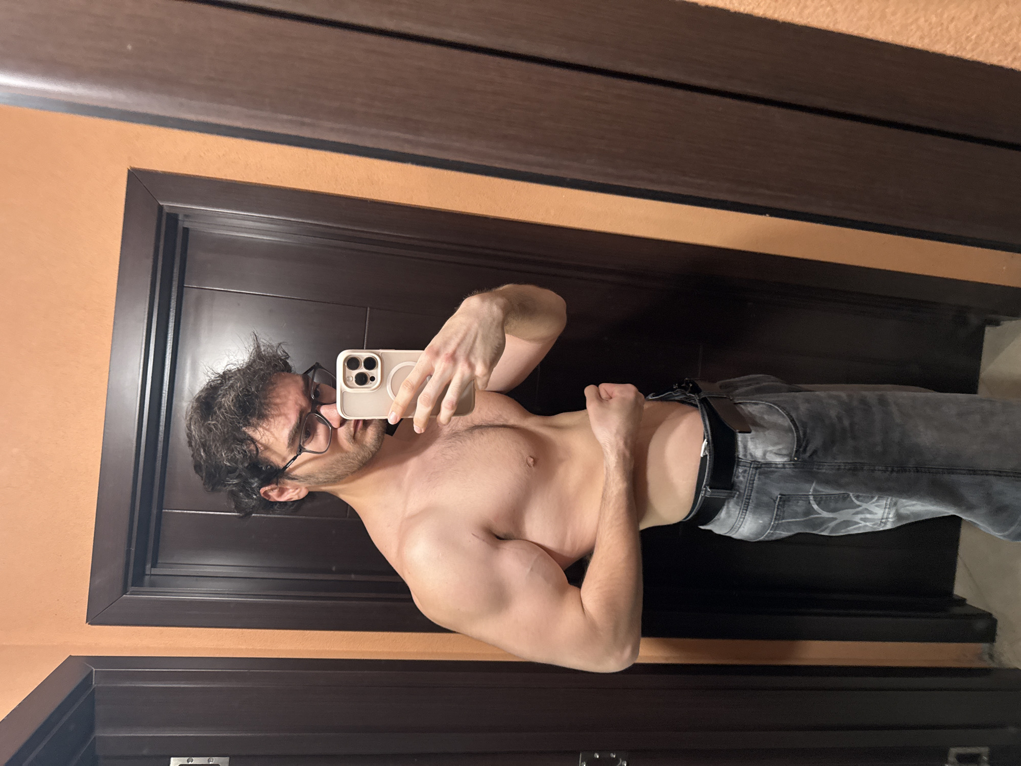 The image captures a mirror selfie of a man indoors, likely in a room in Blagoevgrad, Bulgaria. The man, fair-skinned with dark, curly hair and a short beard, wears dark-rimmed glasses. He is shirtless, displaying a muscular physique, and is dressed in faded gray jeans with a black belt. His left arm is flexed, highlighting his bicep and shoulder. With his right hand, he holds a light-colored smartphone, featuring a three-camera setup and a visible MagSafe ring on the back, towards the mirror to take the photo. He appears to be looking at the phone screen.
The background shows a dark brown door or wardrobe panel framed by matching dark brown trim, set against a light orange wall. The overall lighting suggests an indoor setting with artificial light sources. There is no visible text in the scene.