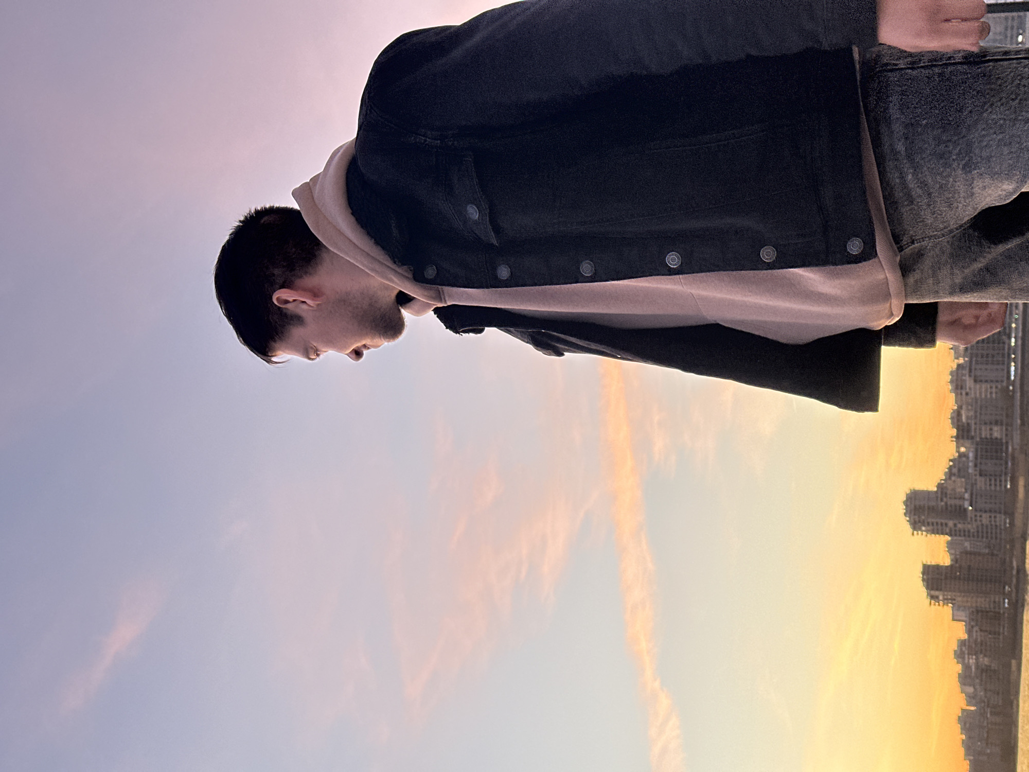 The image captures a young man from a low-angle perspective, positioned against a vibrant sky and city skyline. The original media is rotated 90 degrees clockwise; when viewed correctly (rotated counter-clockwise), the man stands upright, looking downwards towards the city.
The primary subject is a man with short, dark hair and light stubble. He is seen from the upper torso, wearing a dark, possibly denim or corduroy jacket over a light, probably beige or light pink, hooded sweatshirt. His face is in profile, gazing down and slightly to the right with a contemplative or observant expression.
The background is dominated by a sweeping sky, indicating either sunrise or sunset. The sky transitions from a soft lavender and pale purple on the upper left to brilliant oranges and yellows on the lower right, with wispy clouds catching the golden light. Below the man, along the lower edge of the frame, a distinct silhouette of a city skyline is visible against the brightest part of the sky. This urban landscape features numerous high-rise buildings, suggesting a modern city.
The scene conveys a sense of peaceful observation from an elevated viewpoint, overlooking the city of Kazan, Russia, during a golden hour. No specific activity beyond posing and observation is depicted, and there is no visible text.