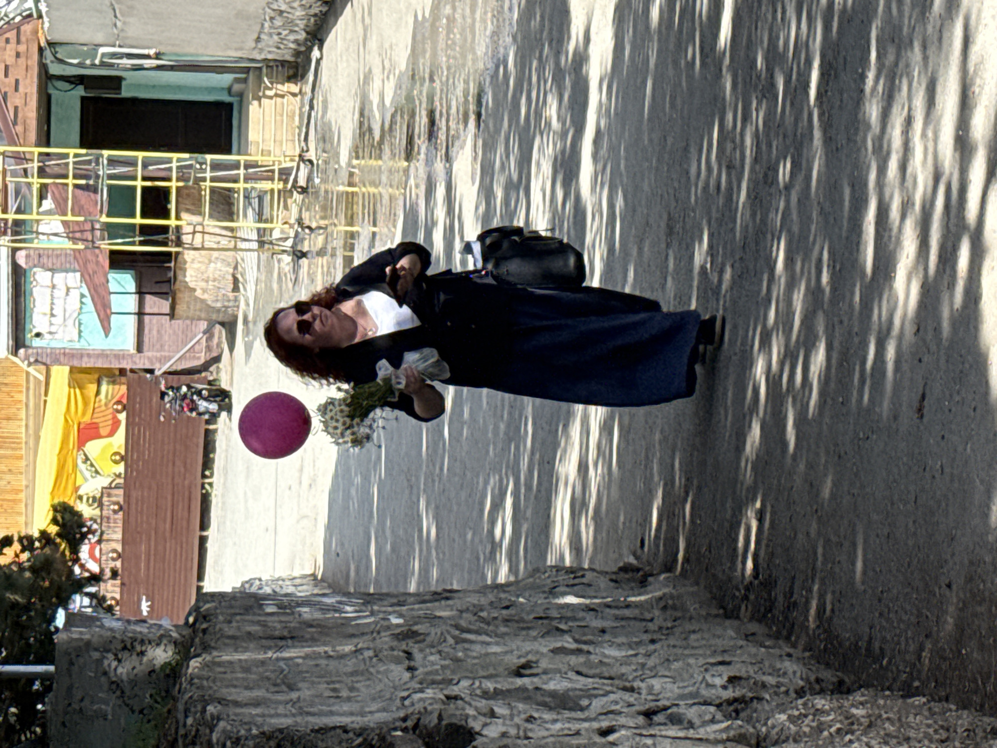 The image shows a woman in an outdoor city setting. She is wearing sunglasses, a black jacket, and a long dark skirt, and she carries a bouquet of flowers and a purple balloon. A black bag is slung over her shoulder. The background includes architectural elements, such as a building with a window and a yellow railing. The ground is made of concrete, with shadows cast across it. The setting is Unknown, Unknown.