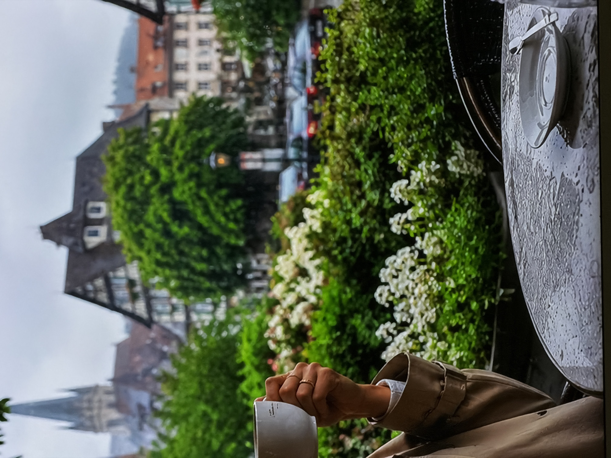 This image, taken in Sofiivska Borshchahivka, Ukraine, presents an outdoor scene from a slightly elevated perspective, possibly from a cafe or balcony. In the immediate foreground, the arm and hand of a person are visible. The person is wearing a light brown or beige trench coat and holds a plain white mug. A gold ring can be seen on one of their fingers. To the right, a portion of a round, textured table surface, which appears wet with small droplets, holds a white saucer with a small spoon resting on it.
The middle ground is dominated by vibrant green foliage and bushes adorned with numerous small white flowers, creating a natural screen. Beyond this greenery, the background reveals an urban landscape under an overcast sky, suggesting a cloudy day, potentially after rainfall. Architectural elements include traditional European-style buildings with varied rooflines, some featuring half-timbered designs. A street with several cars is visible between the buildings and foliage. In the distance, a distinct church steeple adds a historical landmark to the town's silhouette. The overall atmosphere is tranquil and contemplative. No discernible text is visible on any objects within the frame.