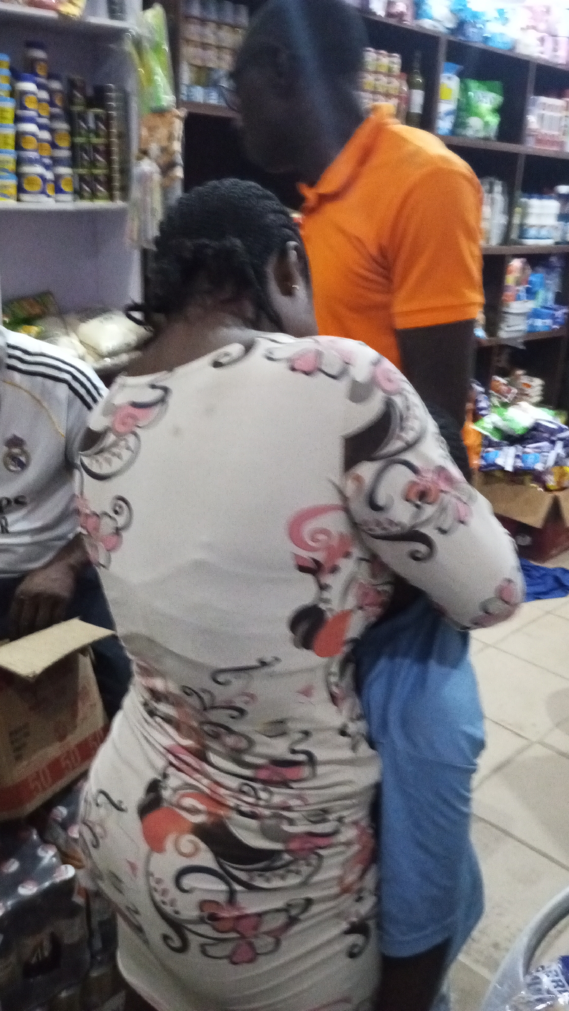 The image shows the interior of a shop in Adankolo, Nigeria. The shop has shelves stocked with various goods. In the foreground, there is a woman with her back to the camera, wearing a tight white dress patterned with pink, gray, and black designs. Next to her on the left is a person wearing a white jersey with black stripes on the sleeves. To her right, there is a man in an orange shirt. A person wearing a blue shirt is obscured by the woman in the white dress. A cardboard box is on the lower left. The floor appears to be tiled.