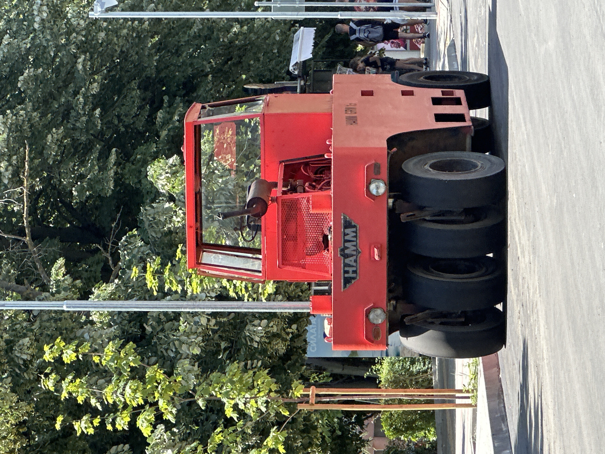 A bright red Hamm GRW 15 roller is parked on a paved surface in Veliko Tarnovo, Bulgaria, likely on a street or construction site, given the presence of trees and a sidewalk. The roller, a heavy piece of construction equipment, has a three-wheel configuration at the front and a single wheel at the rear, with a driver's cabin and visible engine components. The word "HAMM" is prominently displayed on the front of the machine. In the background, several people are visible, including two women and a boy with a backpack, suggesting a public area. The environment is sunny, with lush green trees providing shade. A lamppost stands to the left of the roller, and another utility pole is visible on the right. A sidewalk with a curb separates the paved area from a grassy area with small trees.