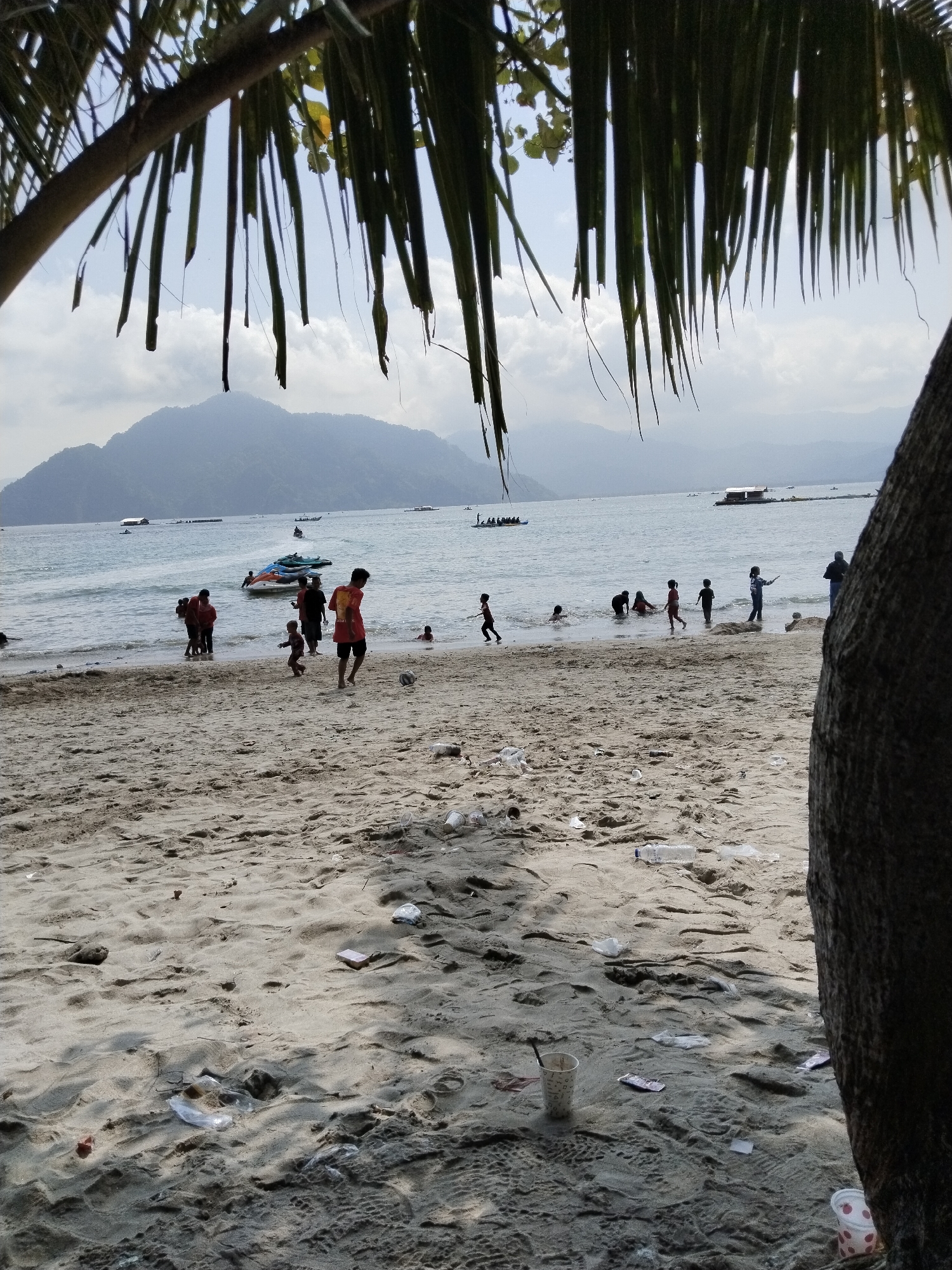 The image depicts a crowded beach in Prigi, Indonesia, on a bright, hazy day. A foreground dominated by sandy beach is strewn with scattered litter, including plastic bottles and wrappers. In the mid-ground, numerous people, predominantly children, engage in various activities near the water's edge. Several individuals are wading or playing in the shallow water, while others are on the sand. One boy is kicking a soccer ball, and a group of adults stand further back. The background features a calm ocean with several boats and jet skis visible. In the distance, a large, verdant mountain range forms the horizon under a cloudy sky. Palm fronds hang into the frame from the top left, and the trunk of a large tree is visible on the right, partially obscuring the scene.