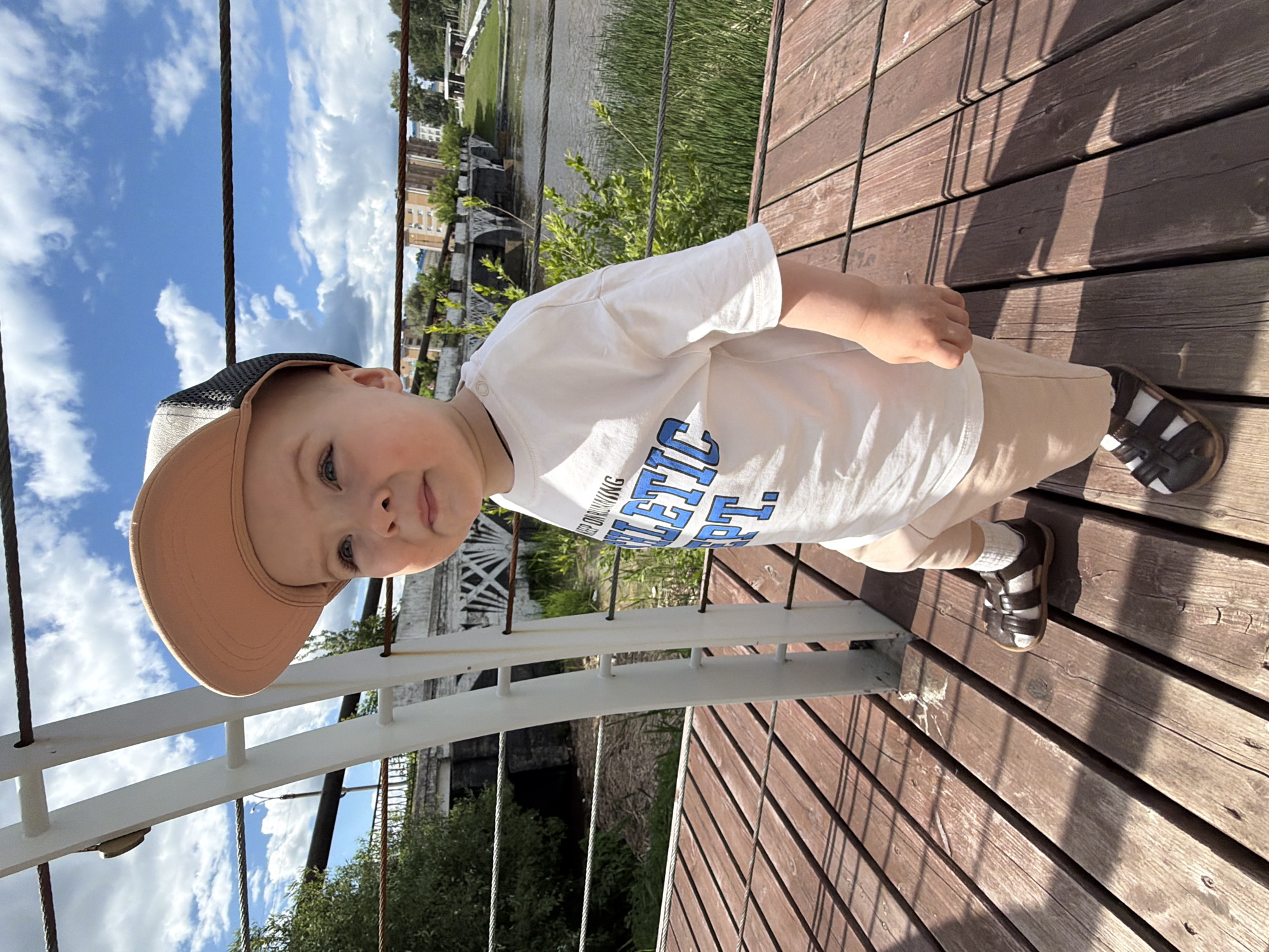In Votkinsk, Russia, on a sunny day with a partly cloudy sky, a young child wearing a white t-shirt with blue lettering that reads "ATHLETIC DEPT.", beige shorts, white socks, and brown sandals stands on a wooden boardwalk. The child is also wearing a light brown baseball cap. The boardwalk is positioned near a body of water, possibly a river or canal, with green reeds growing along the edge. In the background, there is a bridge with white architectural elements and buildings in the distance. A white railing with thick ropes runs along the boardwalk. The child appears to be looking towards the right of the frame.