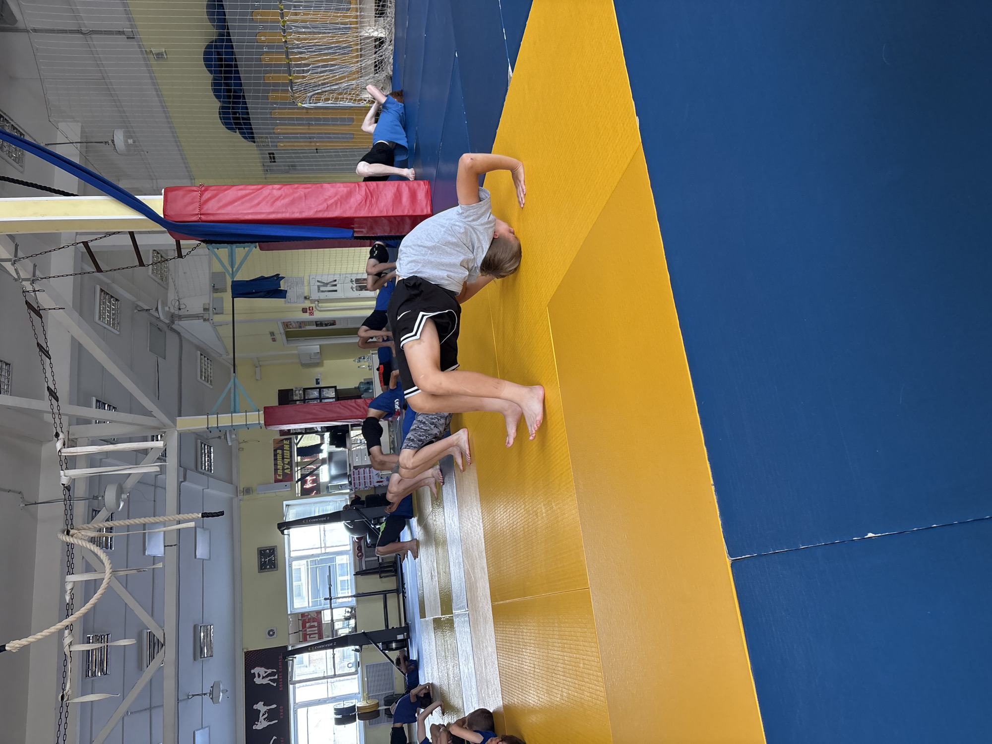 The image depicts a group of young males engaged in gymnastic exercises on a blue and yellow mat floor within a gymnasium. In the foreground, a boy in a grey t-shirt and black shorts is performing a bridge pose, with his body arched and supported by his hands and feet. Several other boys are visible in various stages of similar exercises, lying on the mats in relaxed positions or attempting the same arched pose. The gymnasium environment is indicated by the presence of overhead ropes, chains, lighting fixtures, and a net enclosure in the background. There are also weightlifting machines, a clock, and posters visible on the walls. The overall scene suggests a training session or class for young athletes.