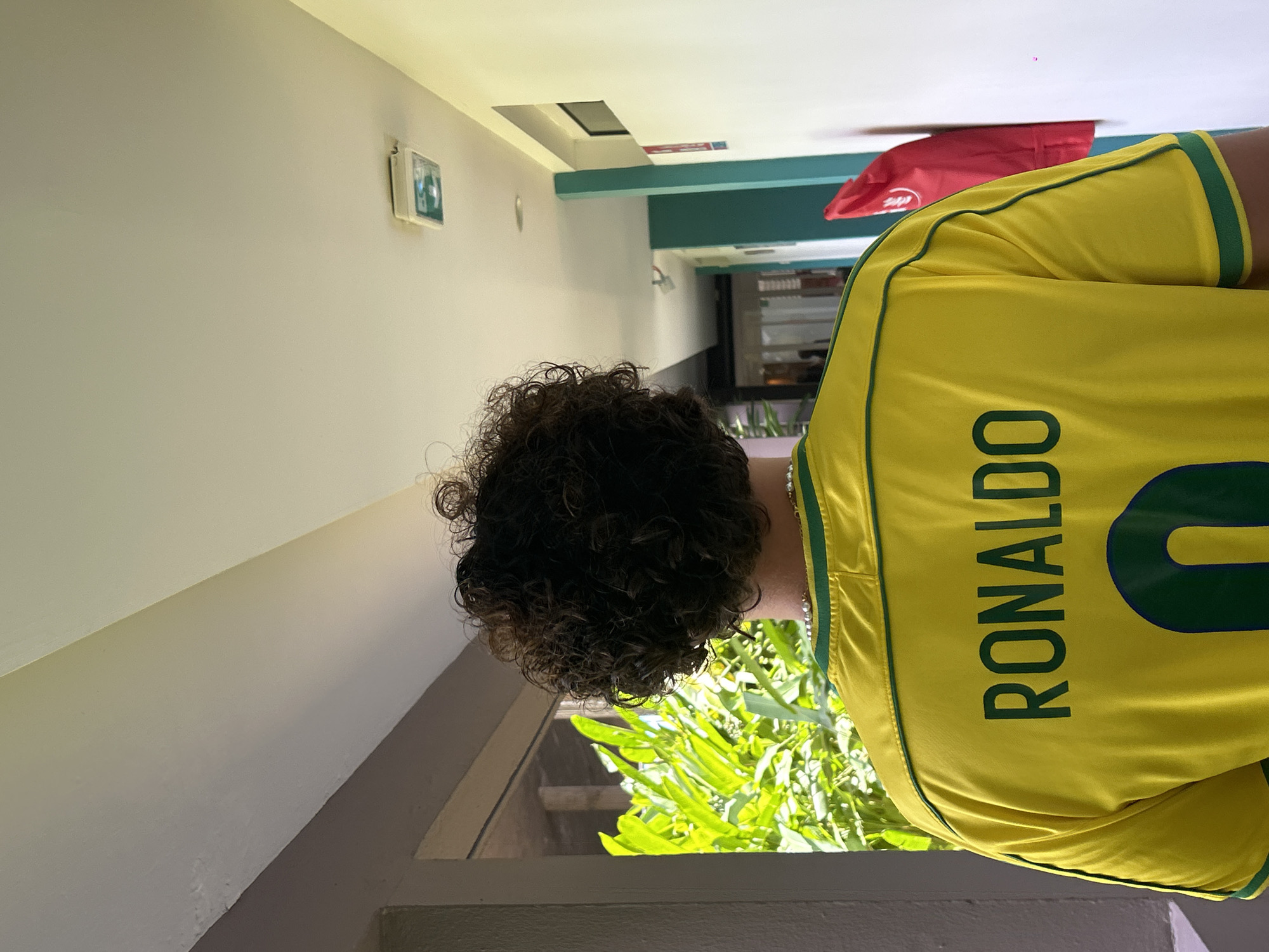 A person wearing a yellow soccer jersey with the name "RONALDO" and the number "0" on the back stands in what appears to be a hallway. The person has curly dark hair. The hallway has a neutral-colored wall on the left and a view of greenery outside a window. A red object is visible further down the hallway. It is Sainte-Anne, France.