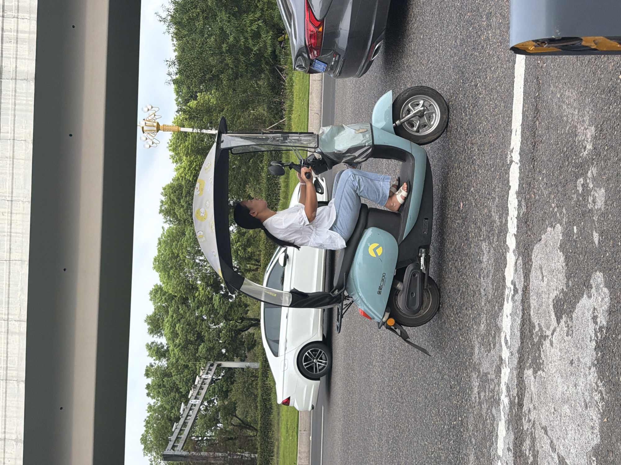 In the city of Jiangjin, China, a woman is riding a light blue electric scooter on a paved road. The scooter has a canopy with a yellow duck pattern. The woman is wearing a white t-shirt and blue jeans. She is holding the handlebars and looking ahead. In the background, a white car is driving on the road. There are trees and a bridge in the distance. The sky is overcast.