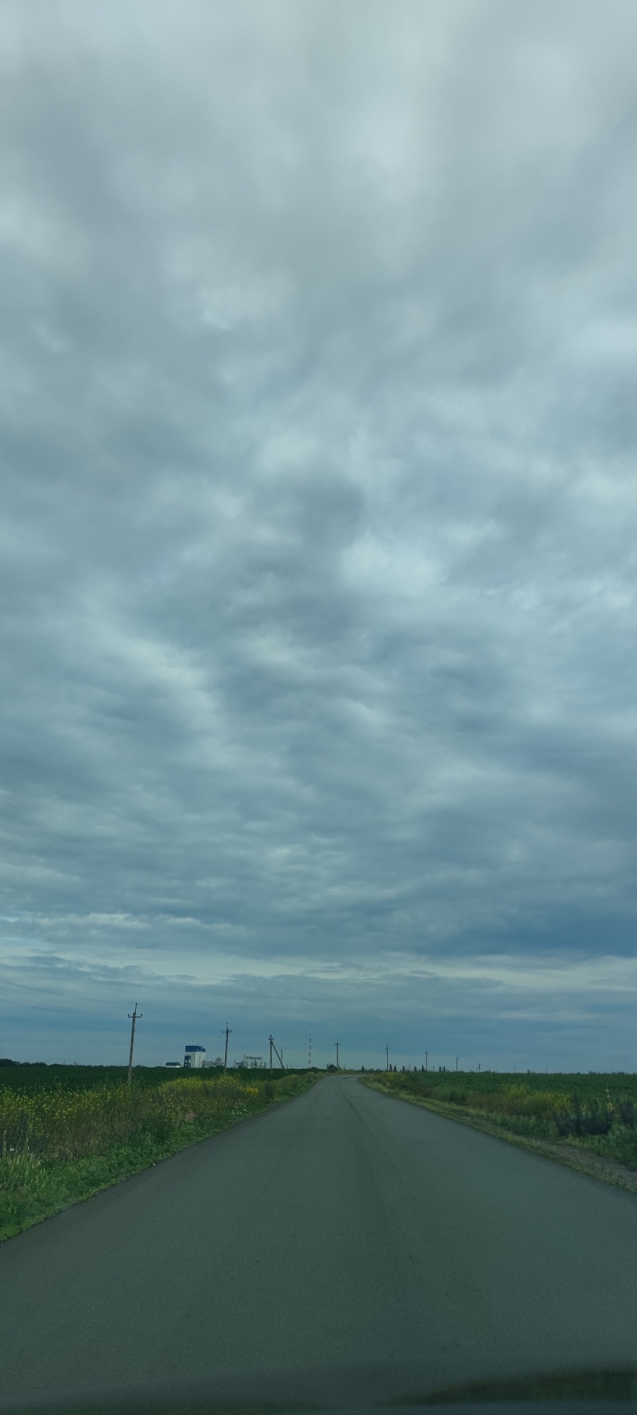 An overcast sky with a dense layer of gray clouds stretches across the frame, filling the upper half of the image. Below the clouds, a two-lane asphalt road recedes into the distance, bisecting a landscape of green fields dotted with yellow flowers. Power lines supported by wooden poles line the sides of the road, and in the far distance, several industrial structures with tall towers are visible, along with more power lines. The overall scene depicts a rural or industrial roadside under a cloudy sky.