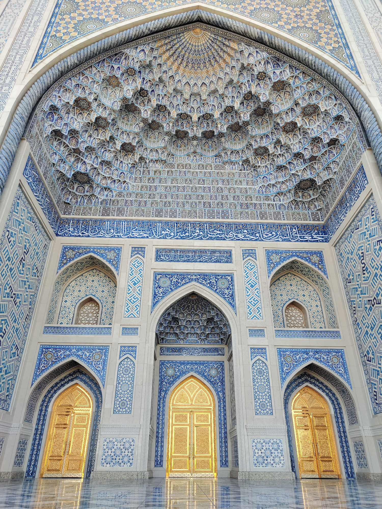 The image shows the grand entrance of the Hazrati Imam complex in Tashkent, Uzbekistan. The scene is a close-up, low-angle shot of the ornate facade of a building, likely a mosque or madrasah within the complex. The architecture is characterized by intricate blue and white tilework, with geometric patterns and Arabic calligraphy adorning the walls and the elaborate muqarnas (honeycomb vaulting) of the main archway. Three large, golden doors, intricately carved and reflecting the ambient light, are visible, suggesting a sense of opulence and grandeur. The floor is polished marble, reflecting the light and the architectural features. The overall impression is one of magnificent Islamic architecture and spiritual significance. No people are visible in the frame.