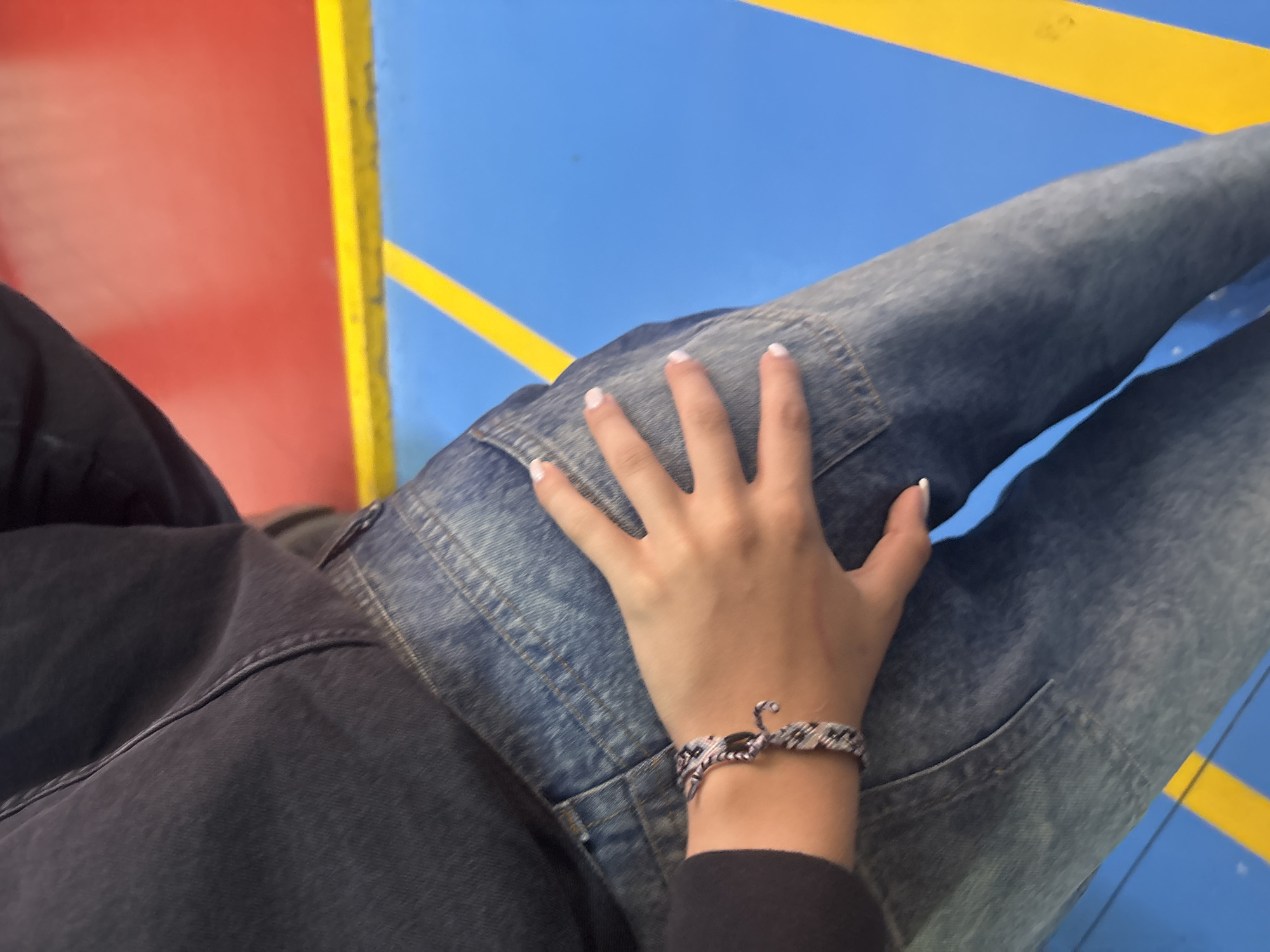 A close-up shot focuses on a person's hand resting on the thigh of another person wearing blue jeans. The hand, with neatly manicured white nails, wears a patterned bracelet with black, white, and pink threads. The background consists of a bright blue surface with a thick yellow stripe and a red surface above it, suggesting an indoor recreational area or play space. The lighting is even, and the image is sharp, with a shallow depth of field that blurs the background slightly.
