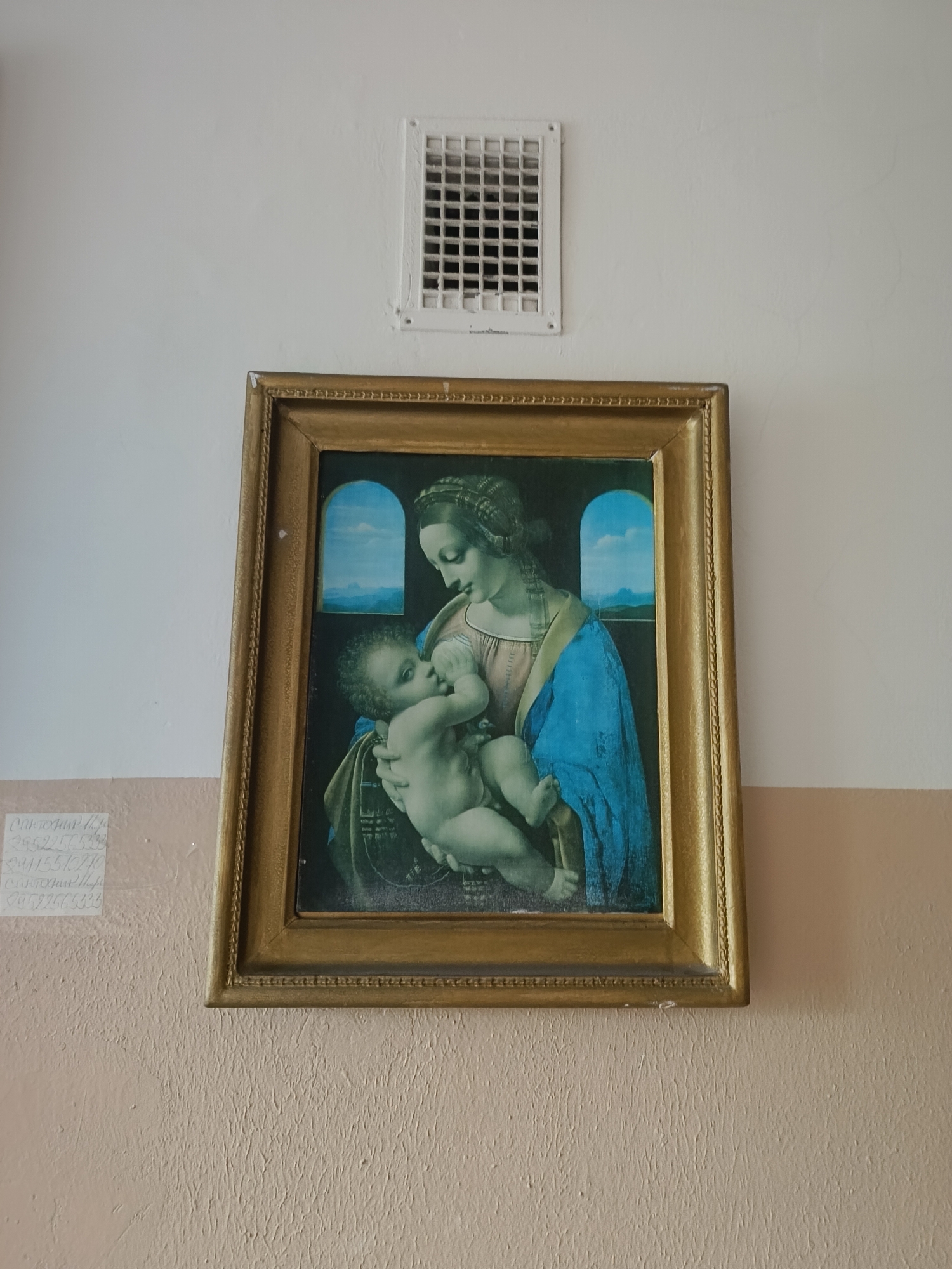 The image displays a framed reproduction of Leonardo da Vinci's painting, "Madonna and Child" (also known as "La Belle Jardinière"). The painting depicts the Virgin Mary breastfeeding the infant Jesus. Mary is shown with a gentle expression, her gaze directed towards the child. Jesus is reaching for his mother's breast. They are positioned against a backdrop that features two arched windows opening to a serene landscape with hills and a blue sky. The painting is housed in a thick, ornate golden frame.
Above the framed artwork, a white air vent is mounted on the wall, which appears to be painted in a light, neutral color. Below the painting, the wall transitions to a textured, light brown surface. To the left of the painting, on the lighter section of the wall, there is handwritten text in what appears to be Cyrillic script. The visible text includes what could be names and numbers. For example, one line appears to read "Святоган Н.Р." followed by numbers like "2952245233" and "2911557090". Another line below seems to be a repetition or a similar entry. The overall setting suggests an indoor location, possibly a room in a building in Arkhangelsk, Russia, given the context provided. There are no obvious cues about the time of day or weather.