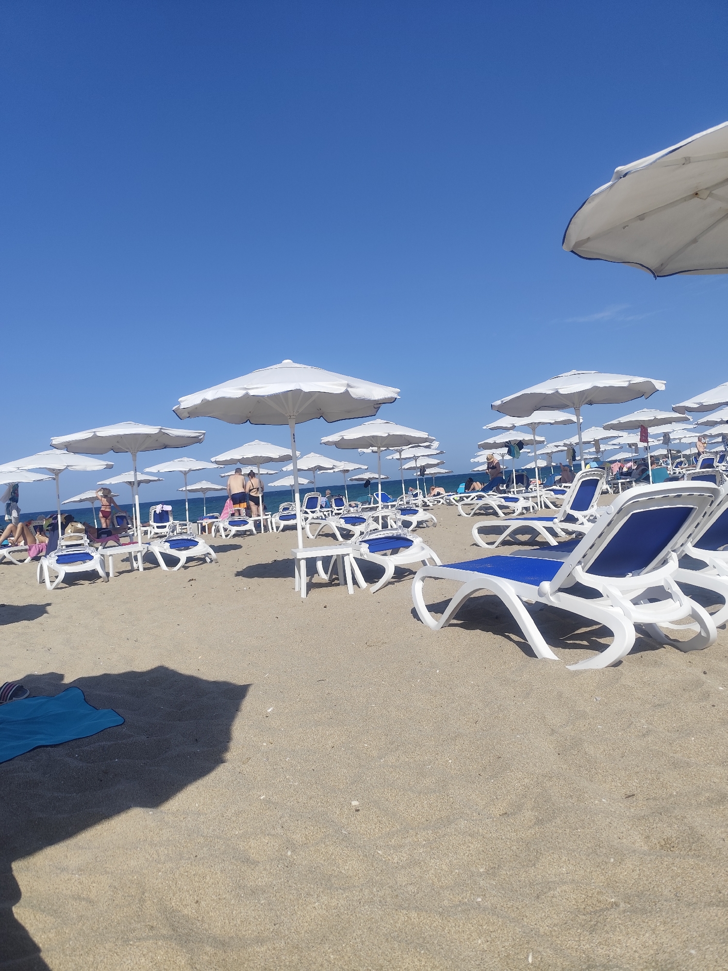 The image depicts a crowded beach scene on a sunny day, likely in Lozenets, Bulgaria, given the context. The foreground is dominated by rows of empty white and blue plastic beach chairs and umbrellas, suggesting a well-maintained tourist area. The sand is light brown and appears fine-grained. In the midground, several people are visible, some sunbathing on chairs, while others stand or walk. A few individuals are dressed in swimwear, further indicating a beach setting. The background shows the calm sea under a clear, bright blue sky. The overall impression is one of a popular holiday destination with ample amenities for beachgoers.