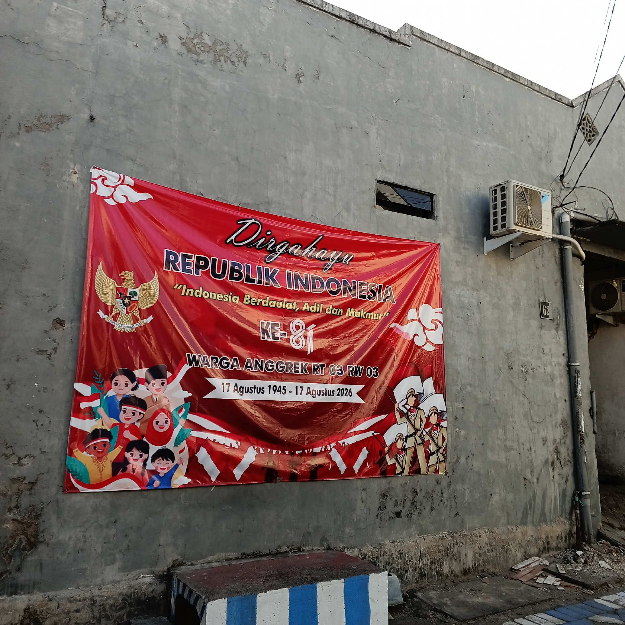 A red banner hangs on a weathered grey concrete wall, depicting an illustration of children and soldiers, likely for a national celebration. The banner reads "Dirgahayu REPUBLIK INDONESIA" (Long live the Republic of Indonesia) in large white letters. Below this, in smaller text, it says "Indonesia Berdaulat, Adil dan Makmur" (Indonesia Sovereign, Just and Prosperous). Further down, it indicates "KE-81" (81st), signifying the anniversary. At the bottom, it reads "WARGA ANGGREK RT 03 RW 03" (Residents of Anggrek RT 03 RW 03), followed by the dates "17 Agustus 1945 - 17 Agustus 2026" (August 17, 1945 - August 17, 2026). The banner features the Indonesian coat of arms, a Garuda bird, on the left side, and stylized clouds at the top. On the right, an illustration shows several figures in military-style uniforms holding flags. The wall itself is rough, with visible cracks and discoloration. An outdoor air conditioning unit is mounted on the wall to the right of the banner, and electrical wires are strung overhead. The ground in front of the wall is rough and appears to be an outdoor area. The scene is likely located in the city of Wedoro, Indonesia.