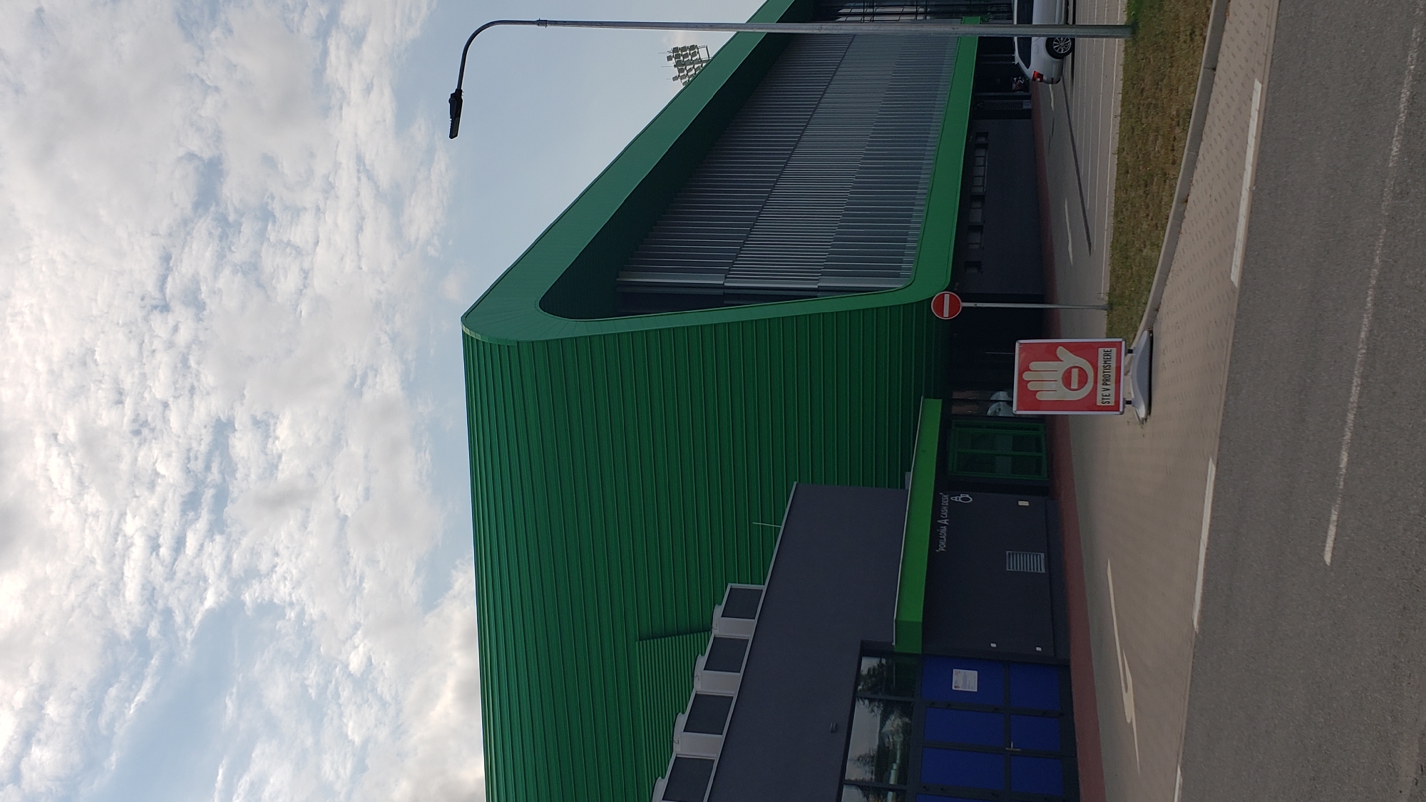 The image shows the exterior of a modern building with a distinctive green, curved roof. The building appears to be a sports or event complex. In the foreground, there is a paved area with road markings and a sidewalk. A portable sign with a red hand symbol and text reading "STE V PROTI SMERE" (You are going the wrong way) is placed on the sidewalk. Further back, there is a smaller dark-colored building with blue doors. A silver car is parked to the right of the main building. The sky is partly cloudy. The building is likely the Arena Prešov in Prešov, Slovakia, based on its architectural style and the presence of sports stadium lighting visible in the background.