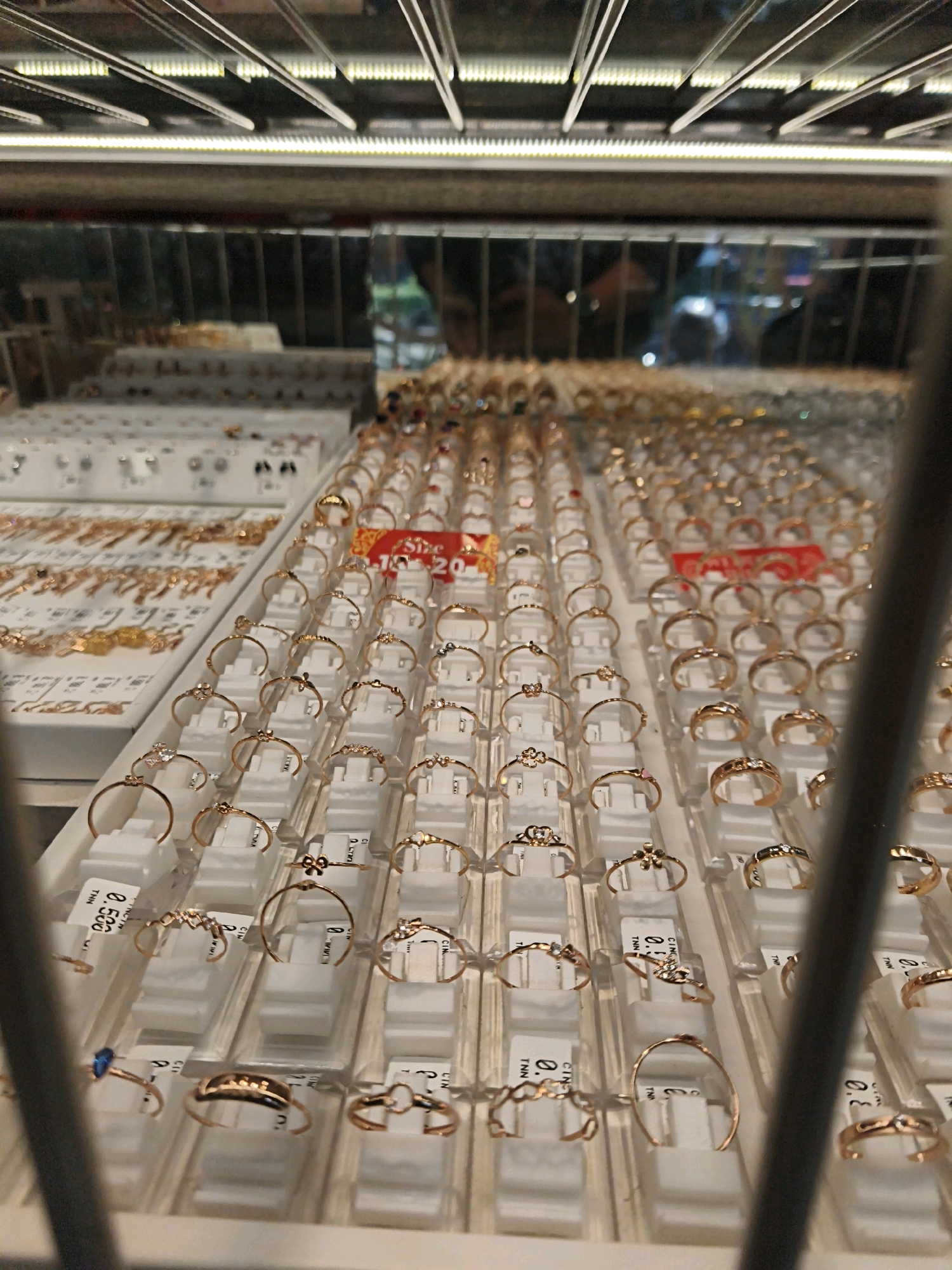 This image shows a display of jewelry, primarily rings, in a store. The rings are arranged in rows on white stands within clear plastic packaging. They appear to be made of gold or a similar colored metal and feature various designs, including simple bands, delicate settings with small stones, and more elaborate decorative elements. Some of the rings have small price tags. In the background, other jewelry items like necklaces and bracelets are visible, also displayed on trays. The lighting suggests an indoor retail environment, possibly a jewelry shop or a department store jewelry counter. Reflections in the glass display case show blurred images of lights and possibly people. The scene suggests a retail setting where jewelry is being offered for sale.
Based on the context of the location provided, this scene is likely located in Surakarta, Indonesia, within a jewelry store or a similar retail establishment.