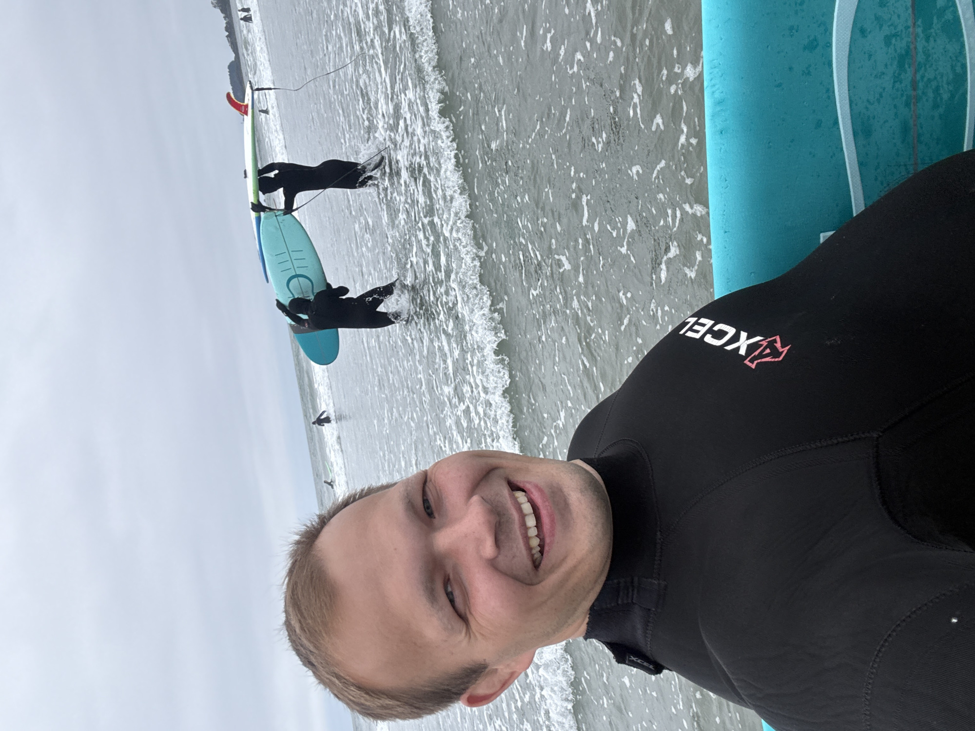 The image depicts a selfie taken at Cox Bay Beach, Tofino, Canada, showing a man in a black Xcel wetsuit smiling at the camera. He is positioned beside a light blue surfboard. In the background, two people in wetsuits stand in the shallow water near the shoreline, one holding a light-colored surfboard with a red fin. The scene takes place on a beach with waves and a gray sky overhead.