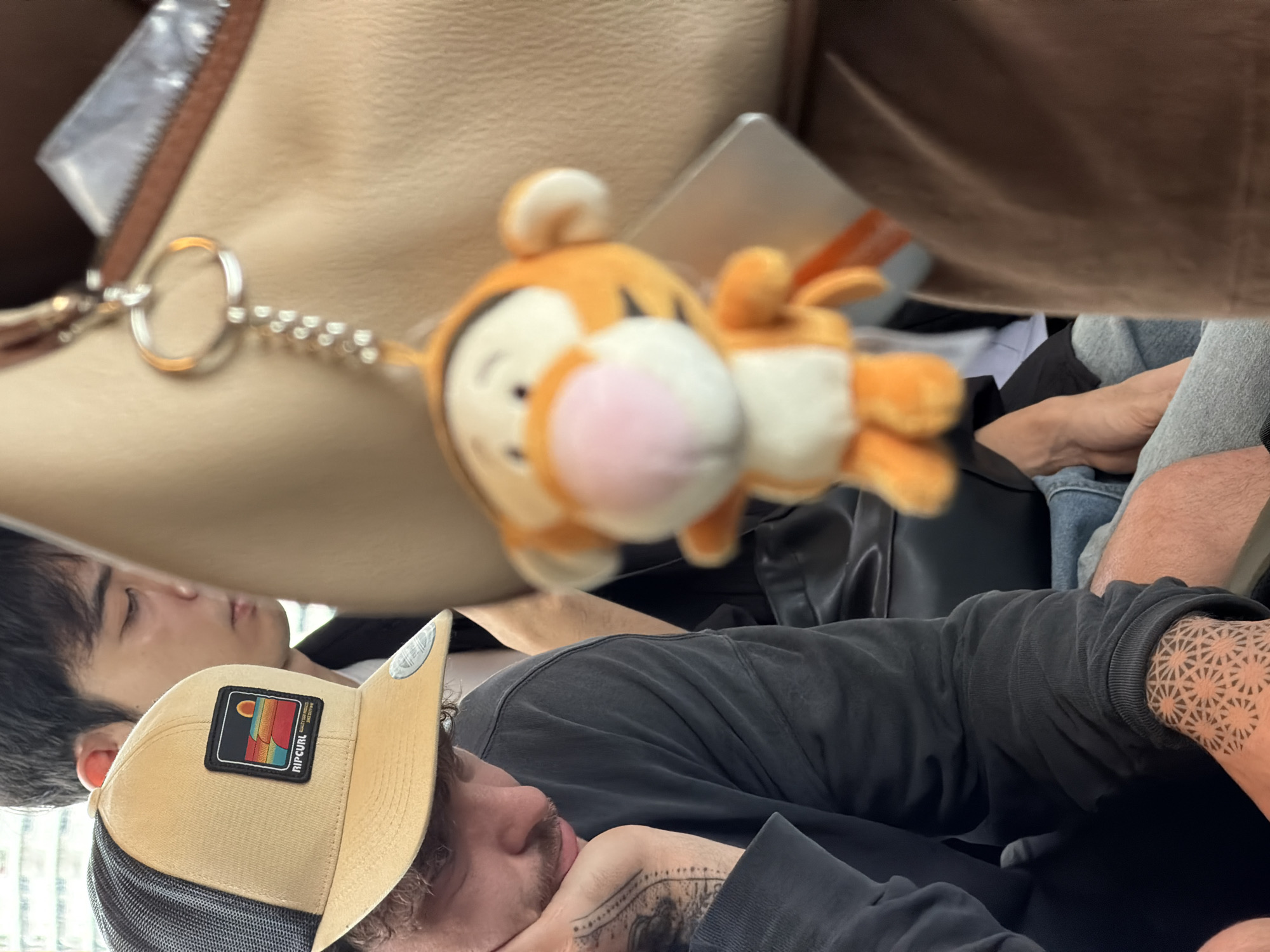 A close-up shot shows a man wearing a tan Ripcurl trucker hat, leaning his head on his hand. He has a mustache and tattoos on his left arm. In the background, another person is visible, partially obscured. A plush Tigger keychain hangs from a metal ring, obscuring the foreground slightly. The scene appears to be indoors, possibly on public transportation, given the presence of seating and the overall composition. The environment suggests a casual setting, with no specific indicators pointing to Bangkok, Thailand.