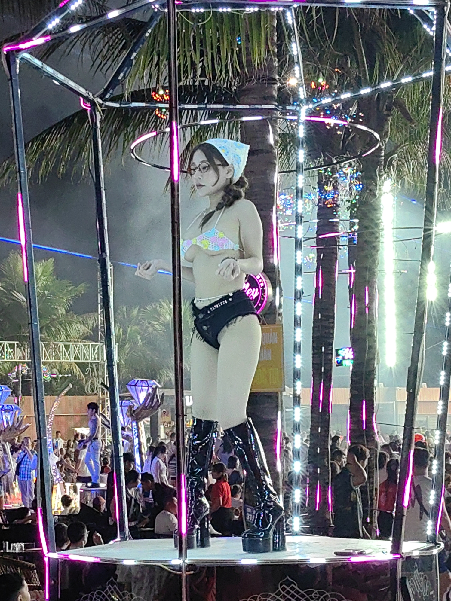 A woman wearing a bikini top, shorts, and knee-high platform boots stands on a platform surrounded by a crowd of people, with illuminated palm trees and structures in the background. The scene is located in Bai Chay Ward, Vietnam.