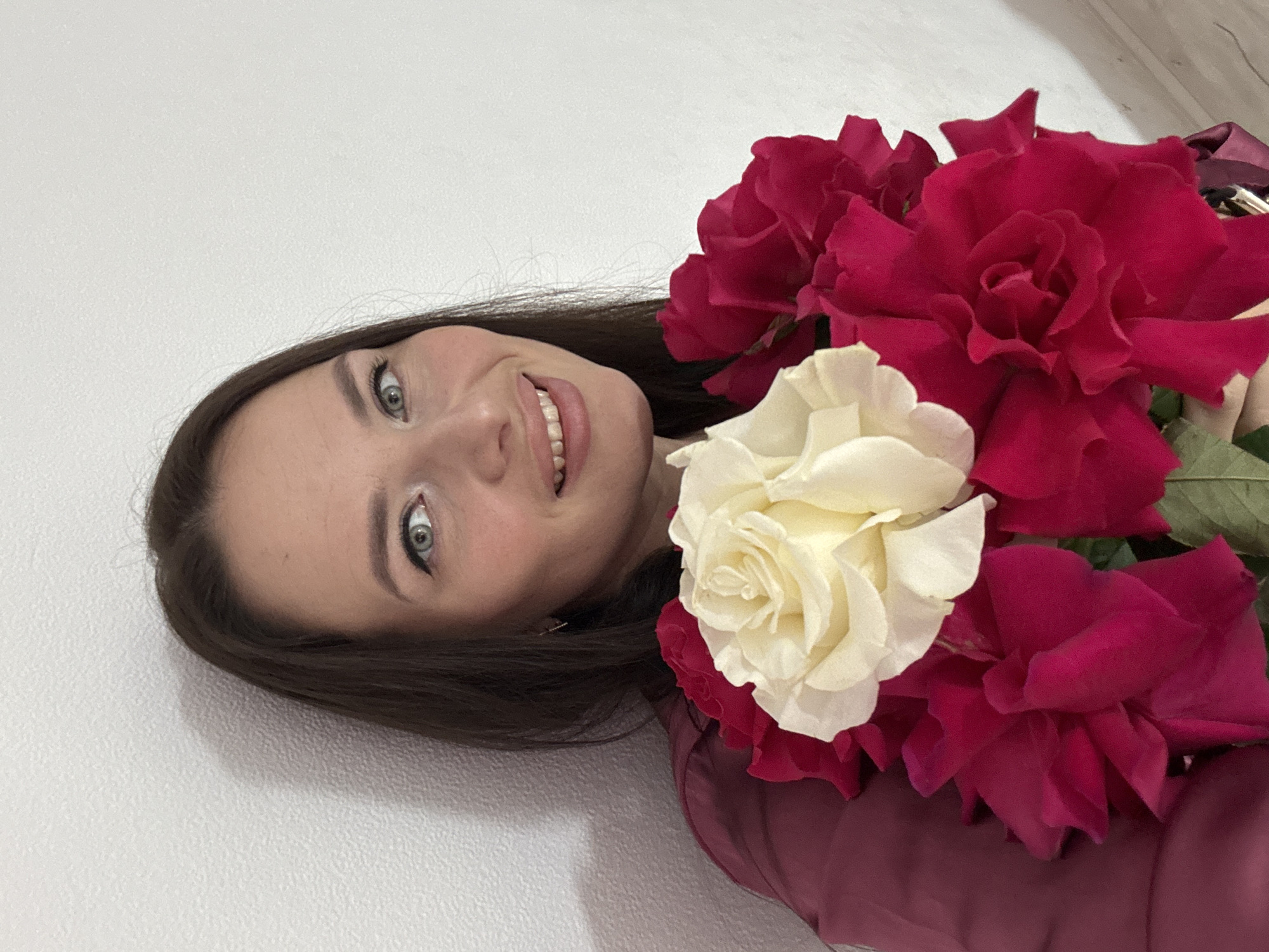 A medium shot shows a young woman with long, dark hair and fair skin, holding a bouquet of red and white roses. She is positioned in the lower left quadrant of the frame, with her face turned slightly towards the camera and a gentle smile. Her eyes are light blue, and she is wearing subtle makeup, including eyeliner and lipstick. The woman is dressed in a dark red blouse or dress. The roses, prominent in the foreground, are large and fully bloomed, with several deep red roses and one creamy white rose. The background is a plain, textured white wall, with a faint shadow cast on it to the left of the woman. The lighting is even, illuminating the woman and the flowers. There is no information in the image to suggest a specific location in the city of Rusalevka, Russia.