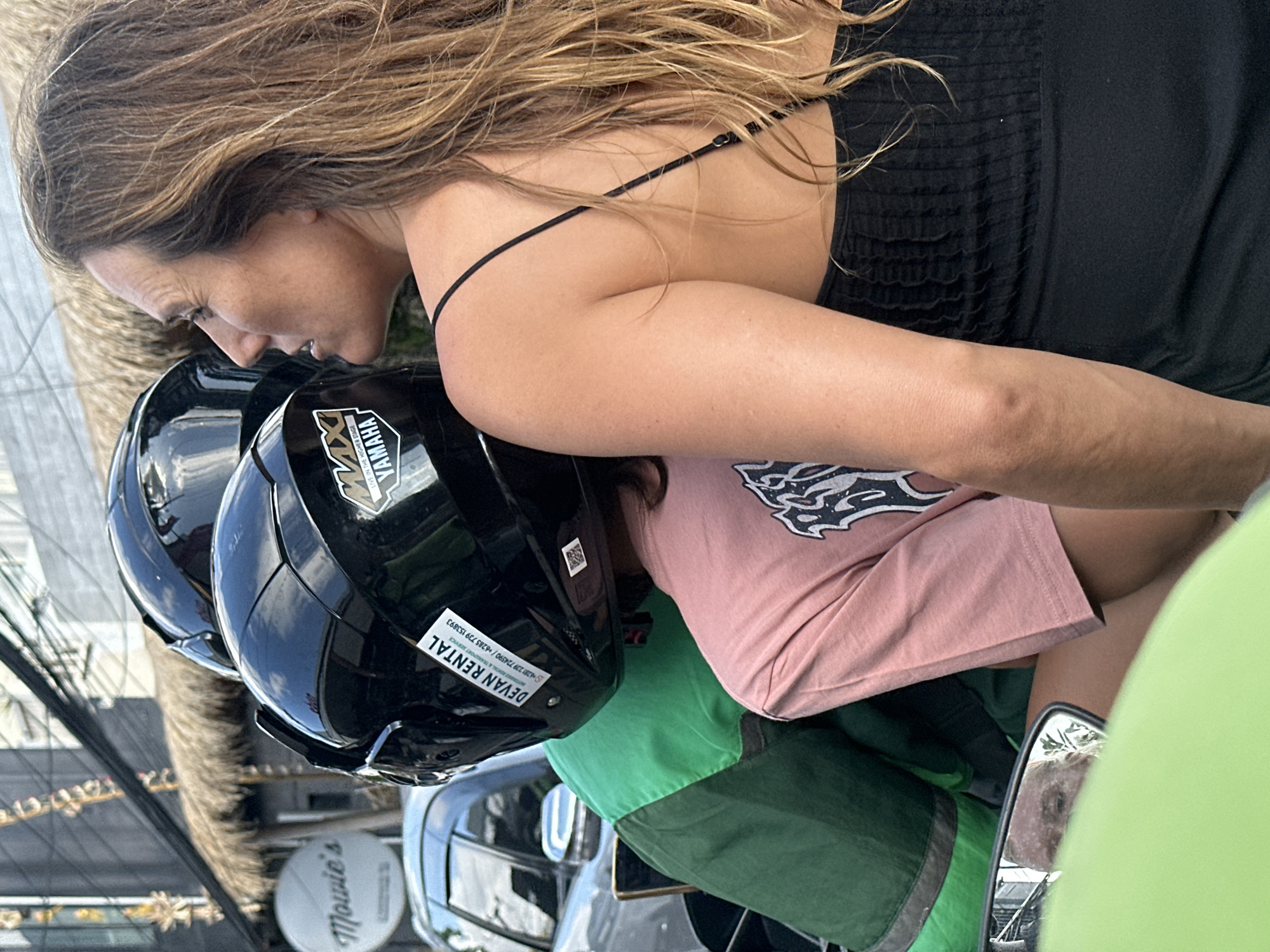 In North Kuta, Indonesia, a medium shot captures a woman from the chest up, her head turned to the right, profile visible. She has long, light brown hair and is wearing a black sleeveless top. In front of her, a person wearing a green jacket and a pink t-shirt is mostly obscured by helmets. Two black helmets are prominently displayed, one with a "Devan Rental" sticker and a "Yamaha" logo. In the background, a building with a thatched roof and a circular sign that reads "Mowie's" is visible. A silver car is parked to the left. The scene appears to be on a street.