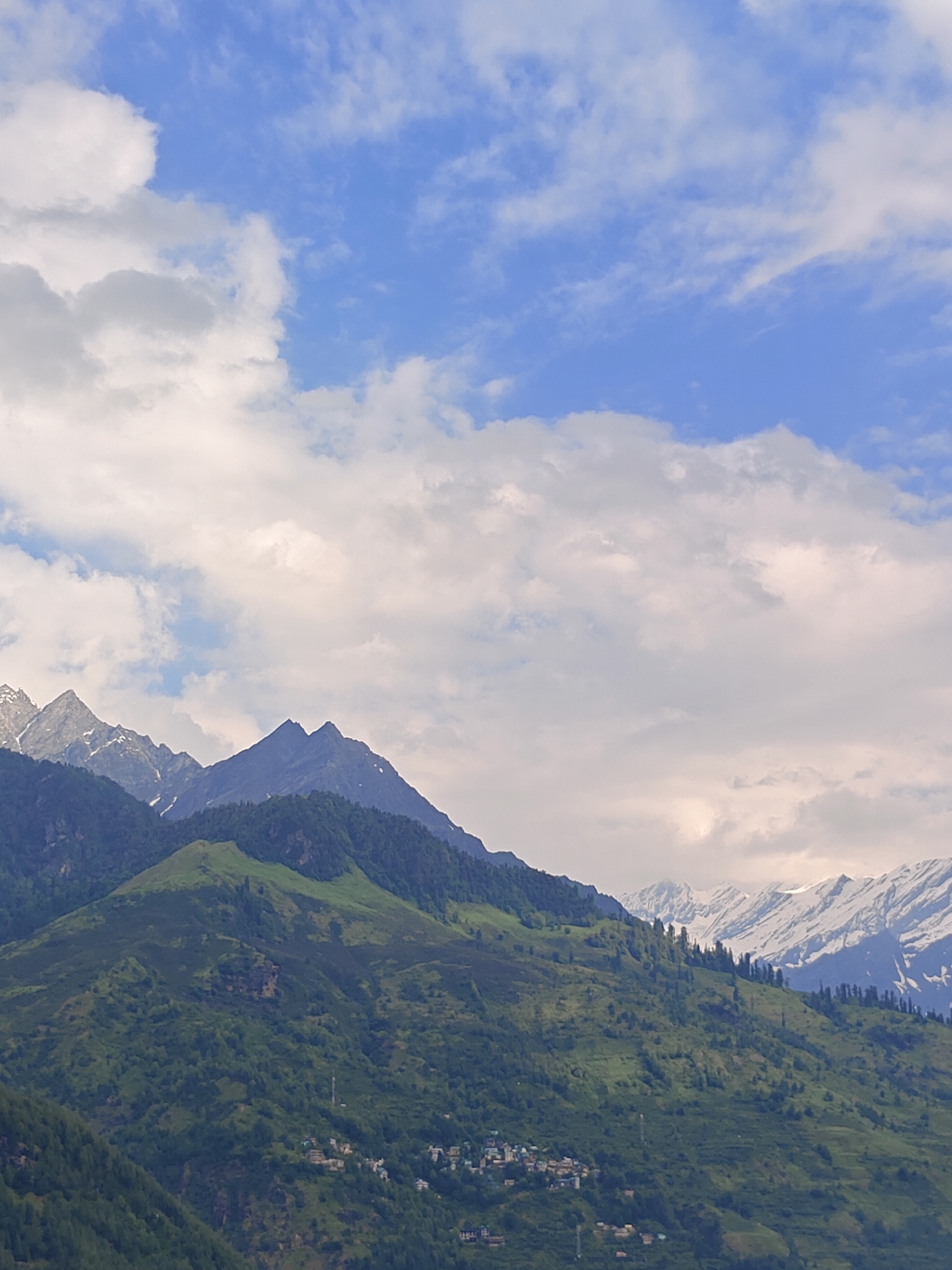 The image shows a panoramic view of the mountains surrounding Vashisht, India. The foreground features lush green hills dotted with trees and small clusters of buildings, suggesting a village or town nestled into the slopes. In the midground, the terrain becomes steeper, with more prominent peaks. The background is dominated by majestic snow-capped mountains, their rugged outlines silhouetted against a partly cloudy sky. The sky itself is a mix of blue patches and white clouds, with some clouds appearing dense and others wispy. The lighting suggests it might be daytime, possibly late morning or early afternoon, with diffused light from behind the clouds. There are no people or animals visible in the image. The overall impression is one of natural beauty and serene mountain landscape.