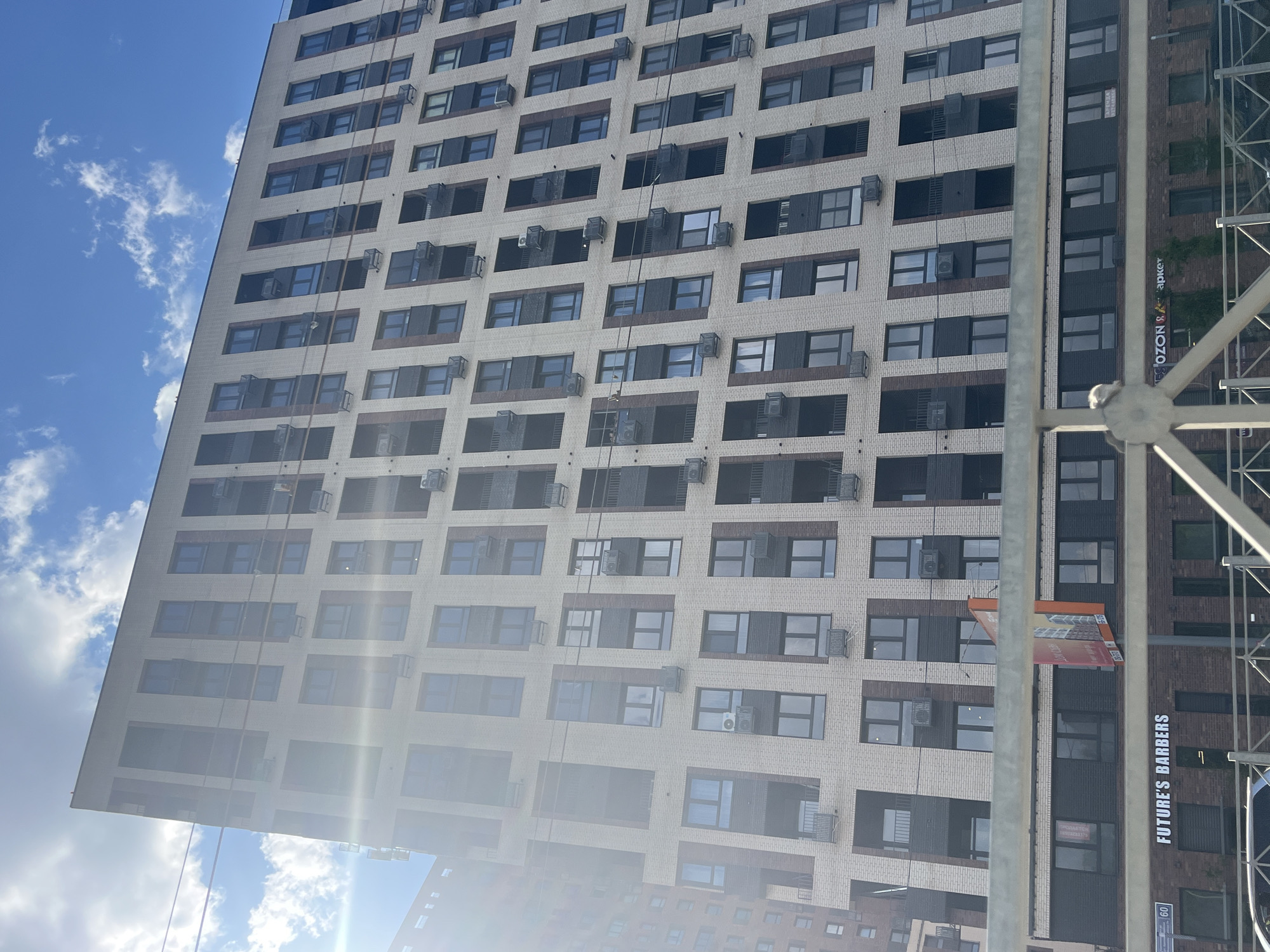 The image shows a modern, multi-story apartment building with a light-colored brick facade and dark window frames. Numerous air conditioning units are mounted on the exterior walls below the windows. In the foreground, a street sign indicates "60", and below that, a storefront sign reads "FUTURE'S BARBERS". Another storefront sign partially visible reads "OZON". The sky is blue with scattered white clouds. The building appears to be located in an urban environment.