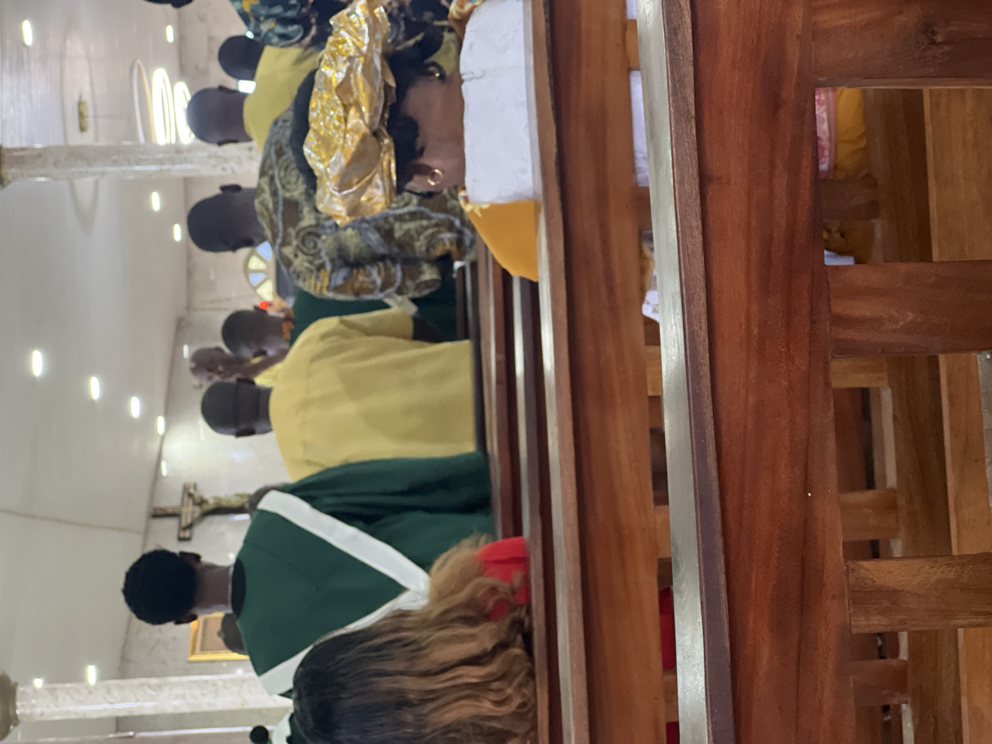 The image depicts the interior of a church, likely in Lagos, Nigeria, focusing on people seated in wooden pews. In the foreground, the back of a woman with long, wavy brown hair and wearing a red garment is visible. She is seated in a pew. Behind her, a woman wearing a white dress and a striking gold headwrap is also seated. Further back, several individuals are seen from behind, facing towards the front of the church. Some are wearing yellow garments, and one person is wearing a green robe with white trim. The church interior features white walls, a high ceiling with lighting fixtures, and a crucifix visible in the background. The lighting is bright, suggesting an indoor setting during daytime. The wooden pews are well-maintained. The overall atmosphere appears to be that of a religious gathering or service.