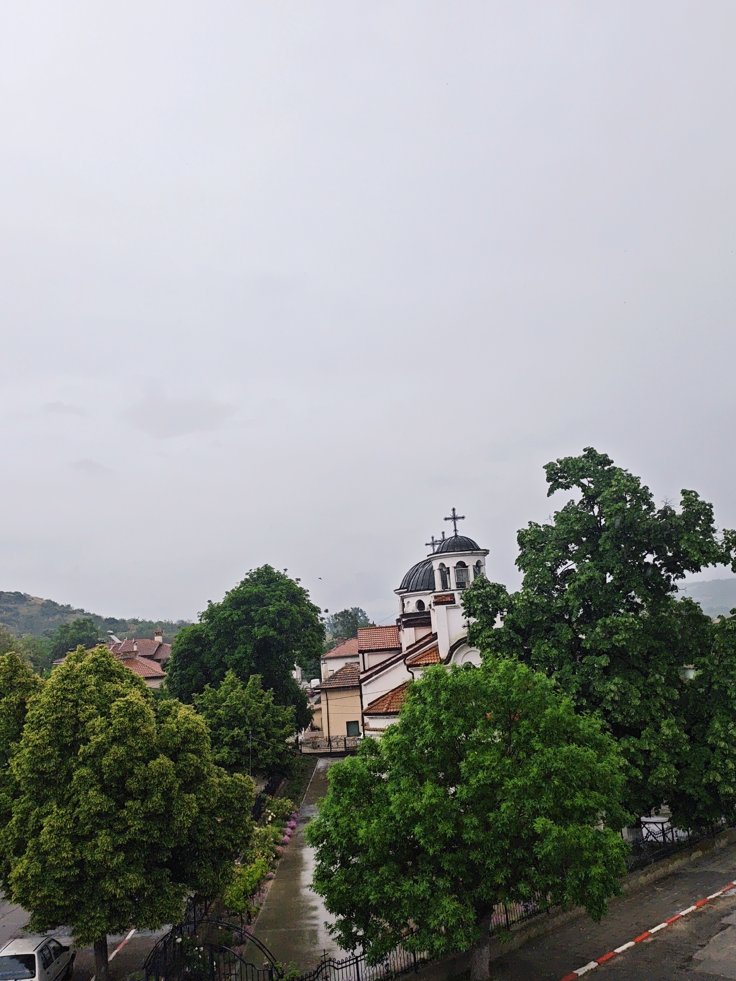 A cloudy day in an urban location of Unknown, Unknown, featuring a church with a dome and cross at the top of its steeple. Several trees surround the church. The roofs of nearby buildings are visible. The image appears to be taken from an elevated position overlooking a street. A car and some sidewalks are partially visible at the bottom of the image.