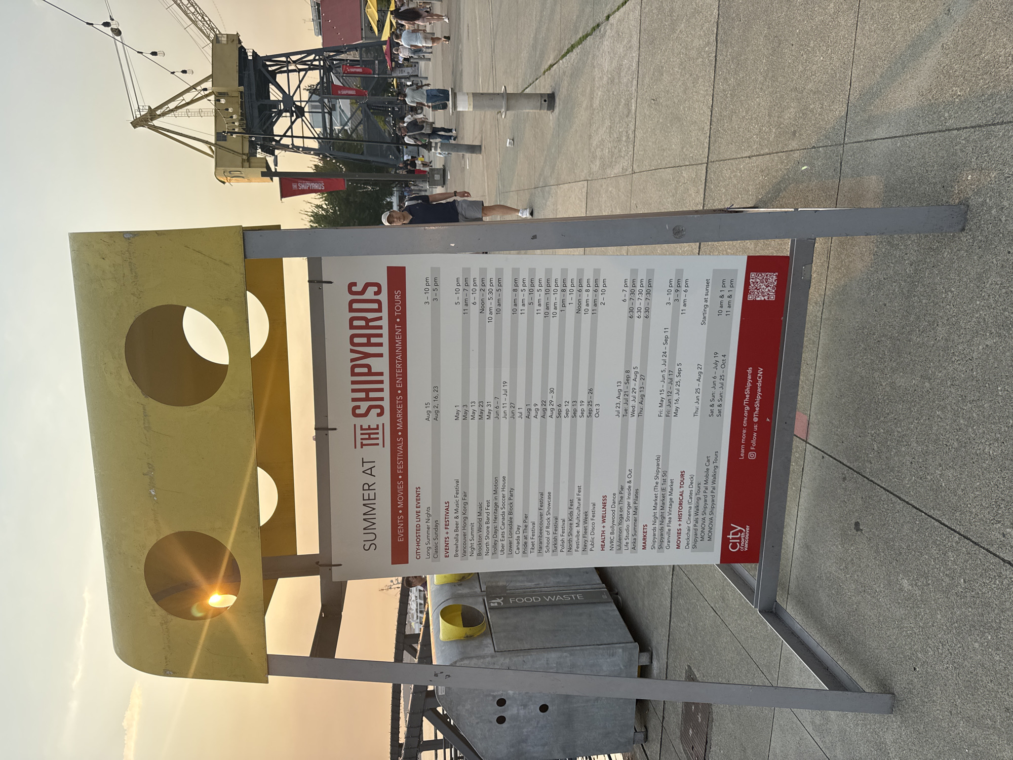 The image shows a large sign for "SUMMER AT THE SHIPYARDS" in the city of North Vancouver, Canada. The sign lists various events, festivals, markets, and activities scheduled for the summer, along with their dates and times. The sign is yellow and white, with red text. In the background, there is a large yellow crane, a body of water, and several people walking around. The sun is setting, casting a warm glow over the scene. The sign is positioned on a concrete walkway, with a metal frame holding it up. There are also some trash and recycling bins next to the sign. A man in a blue shirt and grey shorts is walking past the sign, looking towards the right side of the image.