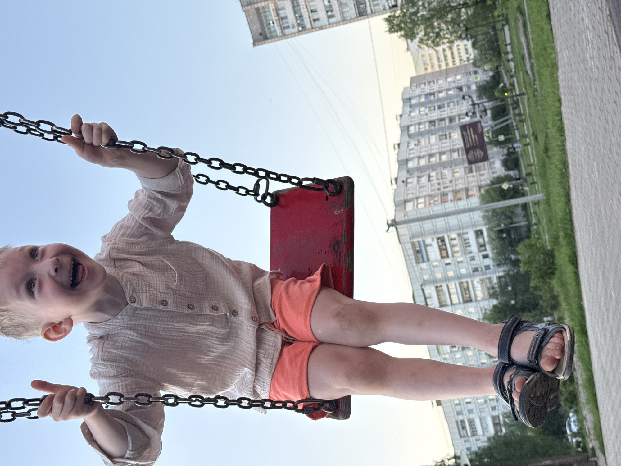 A child is on a swing in what appears to be a park or playground. The child has blond hair and is wearing a light-colored, button-down shirt and orange shorts, and dark sandals. The swing has a red seat attached to chains. The child is holding onto the chains and looks to be laughing or shouting with their mouth open. In the background, there are apartment buildings, greenery, and a concrete path. The location of the city of Chaikovsky, Russia, is not indicated in the image.