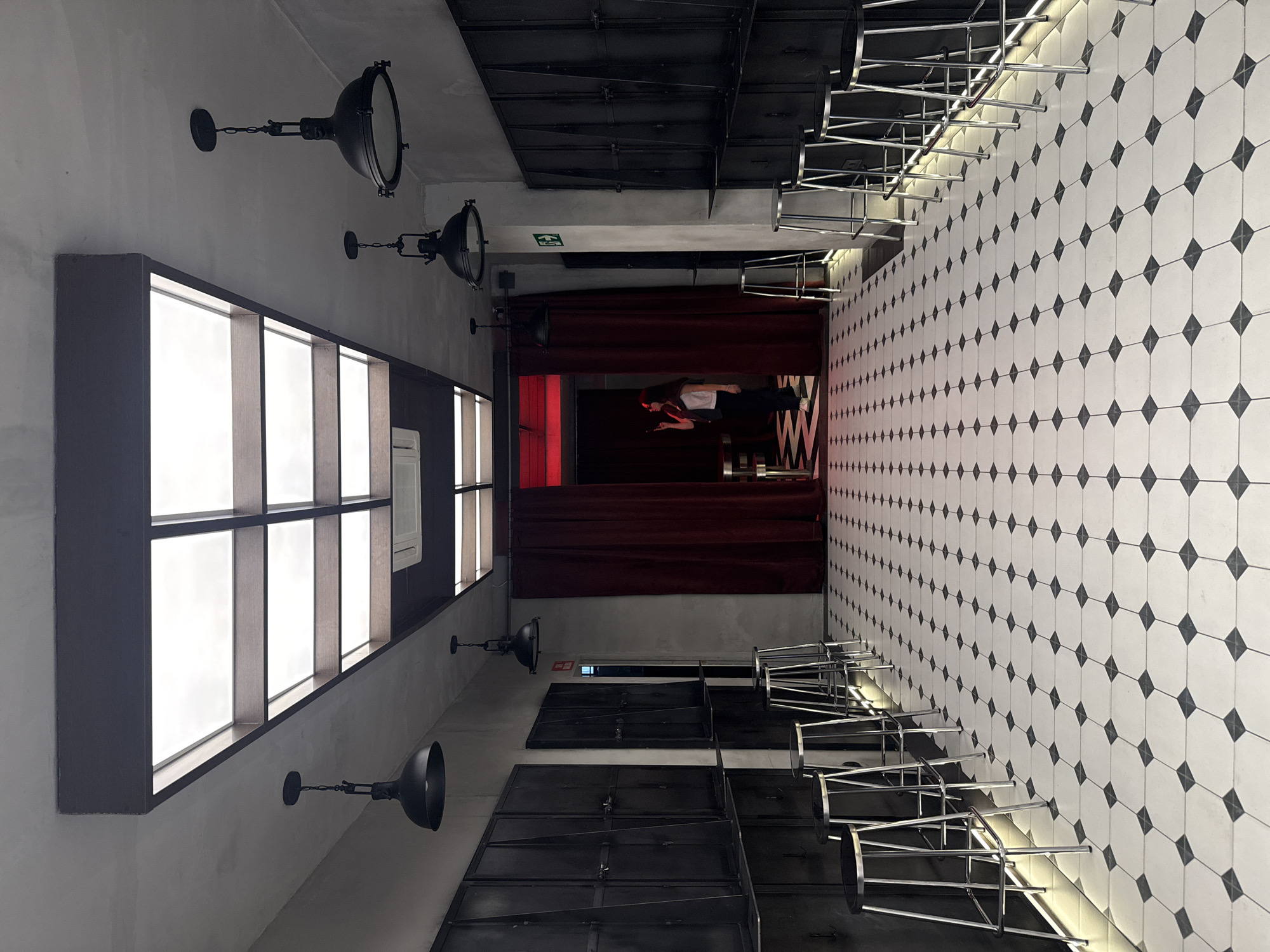 The image depicts the interior of a space, possibly a bar or restaurant, in Madrid, Spain. The location has a modern and industrial aesthetic with exposed architectural elements. Along the left side of the image is a large window with dark frames. Three industrial-style black lamps hang from the ceiling along the left wall.
On the right side, the walls are covered with white tiles patterned with black diamonds. Rows of metal bar stools are neatly stacked along the right wall.
At the end of the space, there is a stage or performance area with red curtains. A person can be seen standing in front of the red curtains.
The ceiling appears to have dark, metal-covered sections.
