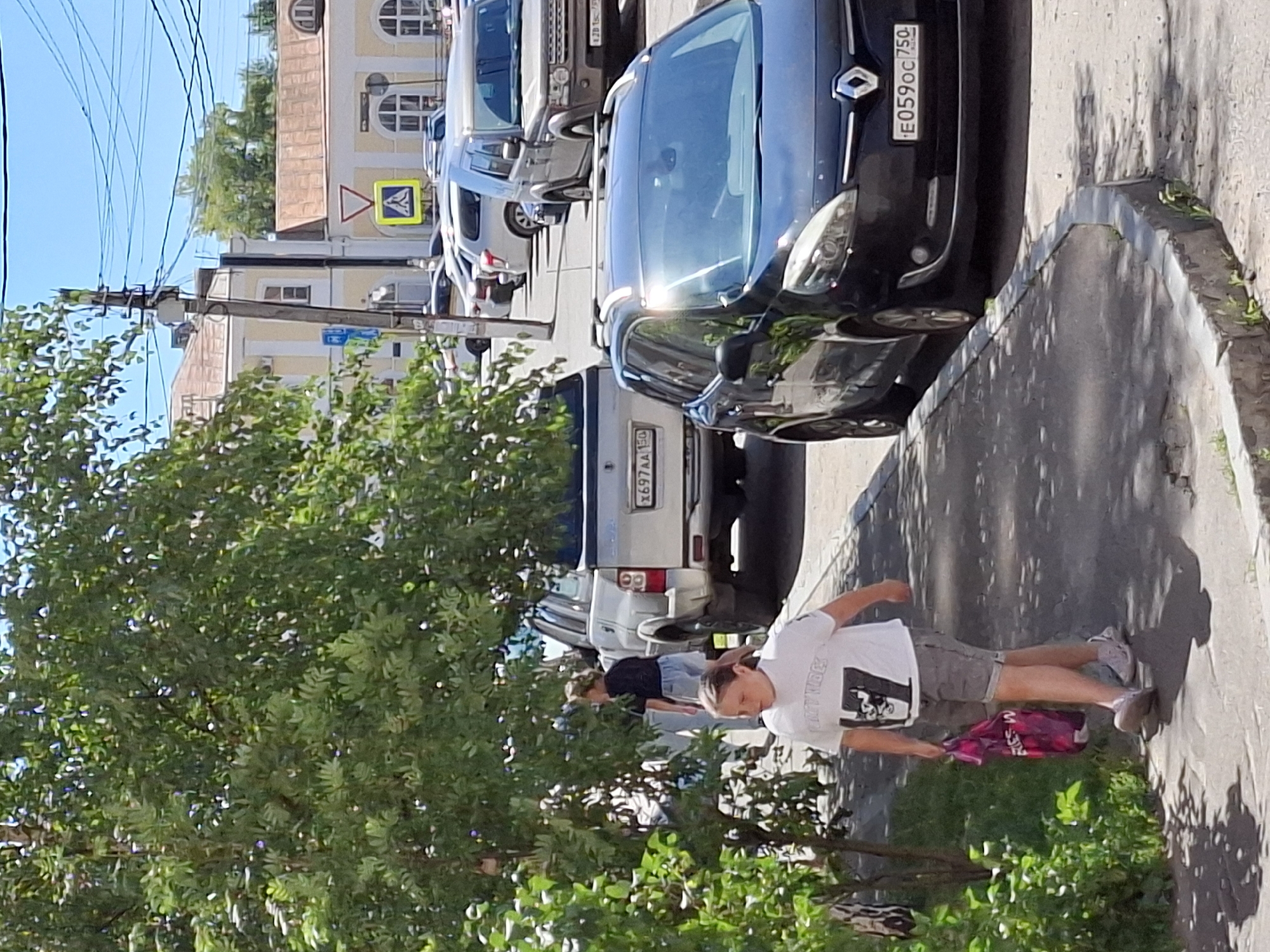 A young person wearing a white t-shirt with a graphic print and grey shorts walks along a paved sidewalk on a sunny day, carrying a pink reusable shopping bag in their left hand. The person is facing forward and slightly to the right. Behind them, a silver SUV is parked, and further down the street, other vehicles are visible, including a black minivan parked on the right side of the road. Trees with green foliage line the sidewalk. In the background, buildings with light-colored facades and tiled roofs are visible. A power pole with electrical wires stands on the left side of the street. A yellow and white pedestrian crossing sign is posted on a pole in the distance.