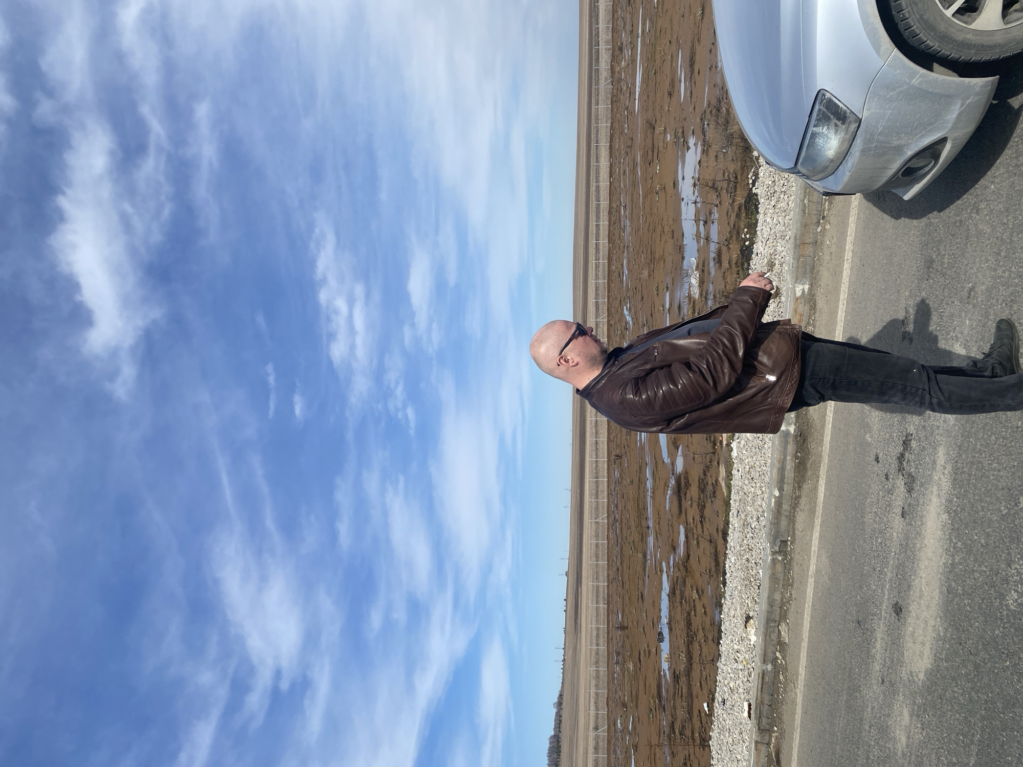 A bald man wearing sunglasses and a brown leather jacket stands on the side of a paved road, next to a silver car. He is looking out over a field with puddles of water. In the distance, a fence line runs across the field. Above, a blue sky is partially covered with white clouds. The location is Kliuchiki, Russia.