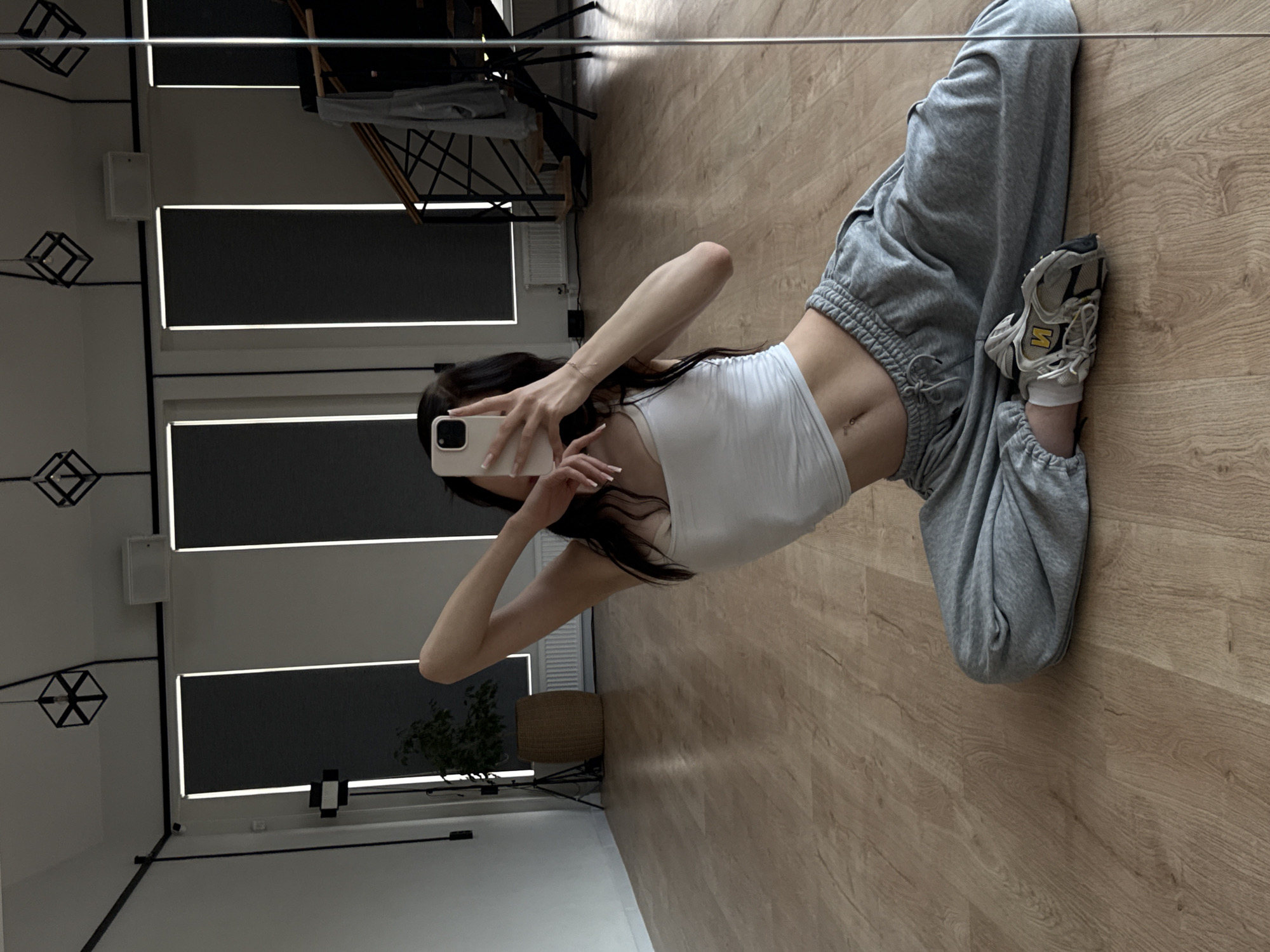 A young woman with long dark hair is sitting on the floor of a dance studio, taking a mirror selfie. She is wearing a white tank top and grey sweatpants, with athletic shoes on her feet. Her left arm is raised behind her head, and her right hand is holding a smartphone, angled to capture her reflection. The studio has a light wood floor, white walls, and large windows with dark blinds. In the background, there is a plant in a woven basket, a metal staircase with clothing draped on it, and modern geometric light fixtures on the ceiling. The overall impression is one of a casual, athletic space.
