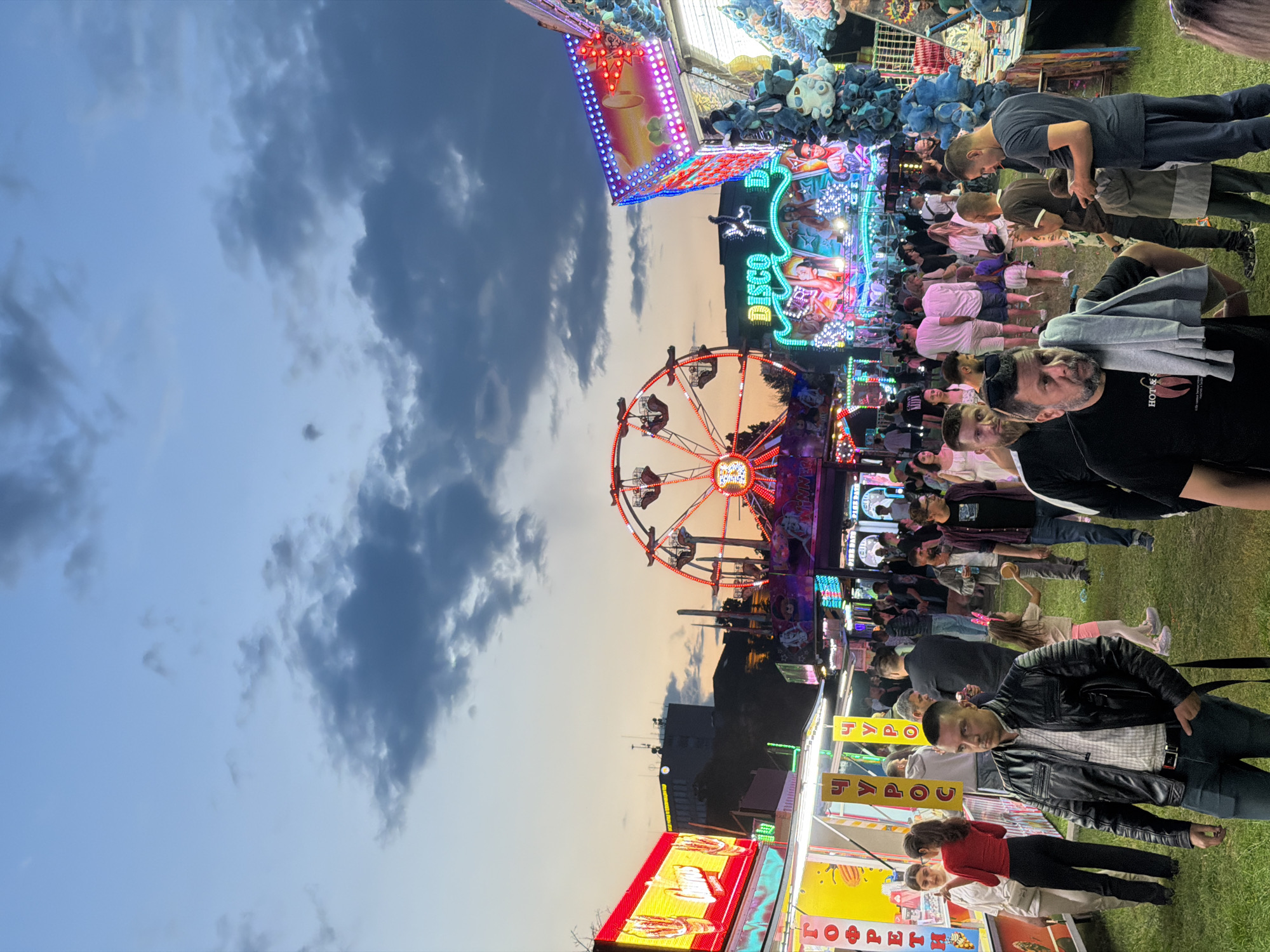 This is a candid, full-frame, medium-angle, vertical photograph of a crowded amusement park at dusk, likely in Sofia, Bulgaria, given the Cyrillic signage visible. The sky is a twilight blue with scattered, dark clouds. In the mid-ground, a Ferris wheel with illuminated cabins is prominent, situated behind various carnival attractions. To the left, a brightly lit food stall displays glowing red signage with white text that appears to read "Cherries." To the right, another attraction, possibly a ride or game booth, is covered in flashing, multicolored lights and adorned with stuffed blue octopuses and other plush toys. The foreground is occupied by a diverse crowd of people of various ages, many of whom are walking, standing, or engaged in conversation. A man in a black leather jacket walks towards the viewer, his hand in his pocket. Another man in a black t-shirt with a graphic print is in the immediate foreground, looking to his left, with a gray sweatshirt draped over his shoulder. The ground is covered with green grass, indicating an outdoor setting. The overall atmosphere is lively and festive, typical of a fairground at twilight.